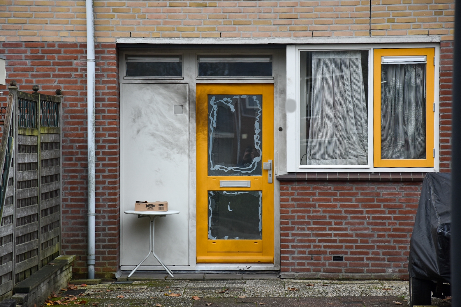 Voordeur van een bakstenen huis met geel kozijn, een klein tafeltje met een kartonnen doos ervoor, en een raam met gordijnen ernaast.