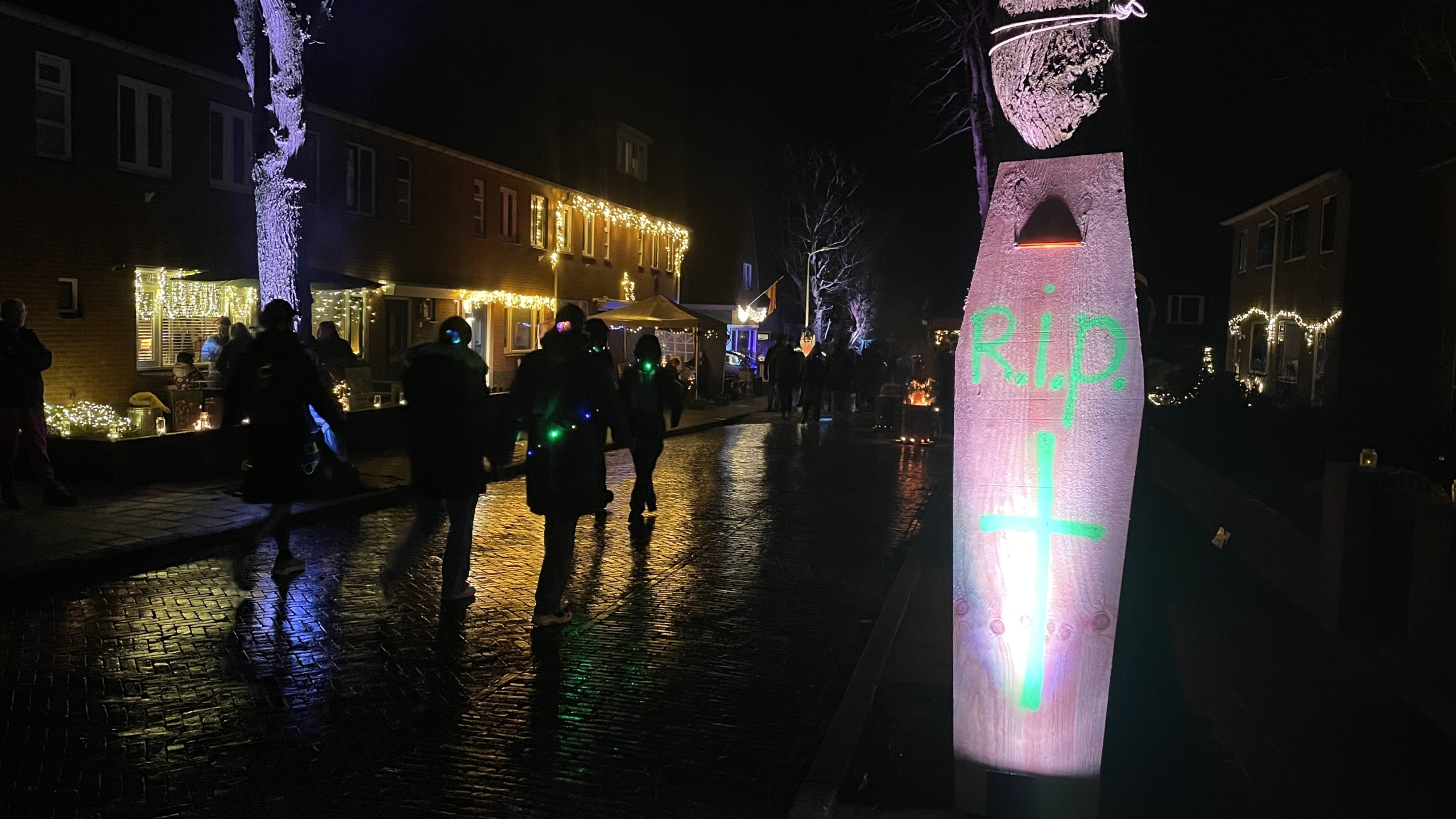 Straatscène 's nachts met een plank waarop "R.I.P." en een kruis in groene verf zijn geschilderd, omlijst door huizen versierd met kerstverlichting.
