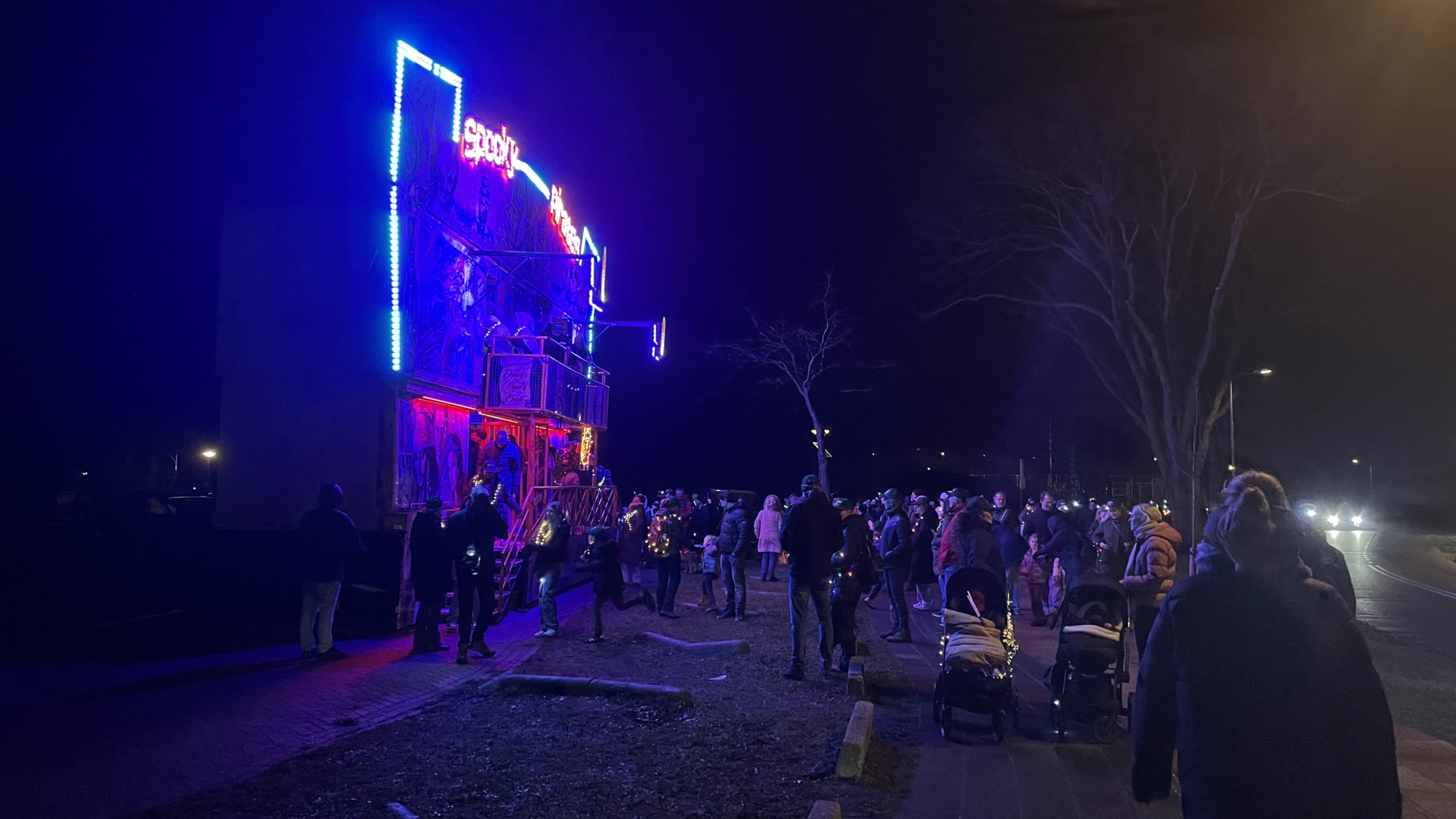 Een groep mensen verzamelt zich 's avonds buiten voor een verlicht attractiegebouw met felle neonkleuren, terwijl sommigen kinderwagens bij zich hebben.