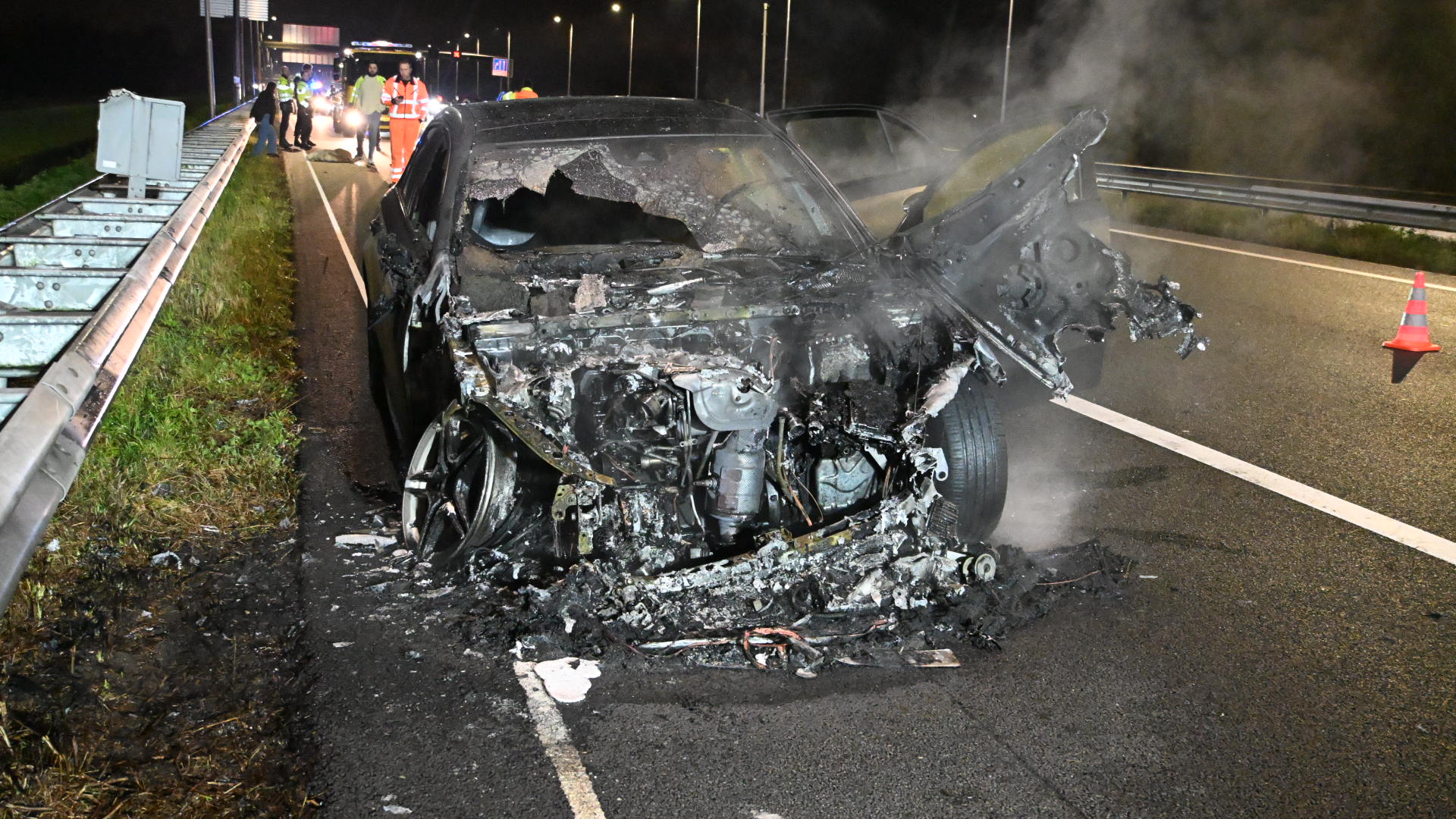 Een auto op de snelweg met ernstige brandschade aan de voorkant en opengeduwd motorcompartiment, omringd door politie- en hulpverleners.