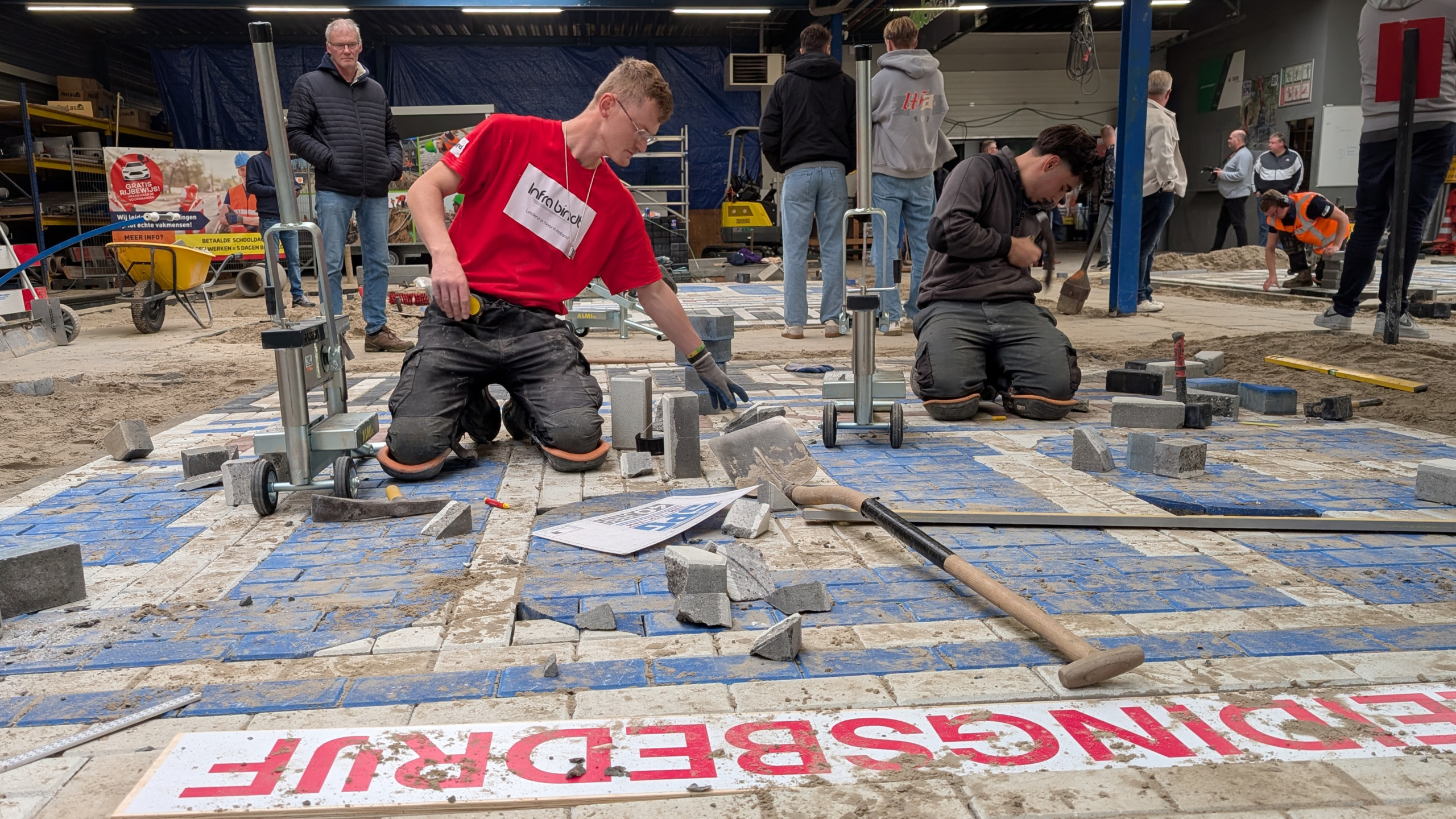 Twee mannen knielen op een bouwplaats binnen een werkplaats en leggen bestrating met stenen en zand. Er liggen gereedschappen zoals een schop en een steenknipper op de grond. Op de achtergrond staan enkele toeschouwers en andere arbeiders.