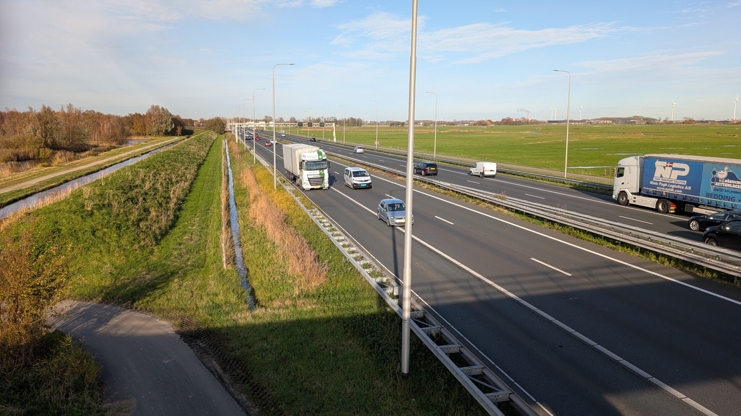 Autos en vrachtwagens rijden over een snelweg omgeven door groene velden, met aan de linkerkant een smal kanaal.