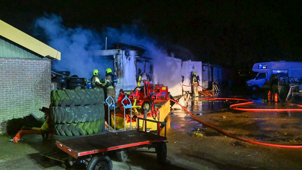 Brandweerlieden bestrijden 's nachts een brand bij een gebouw, omgeven door rook, met een brandslang op een terrein vol met landbouwbanden en apparatuur.