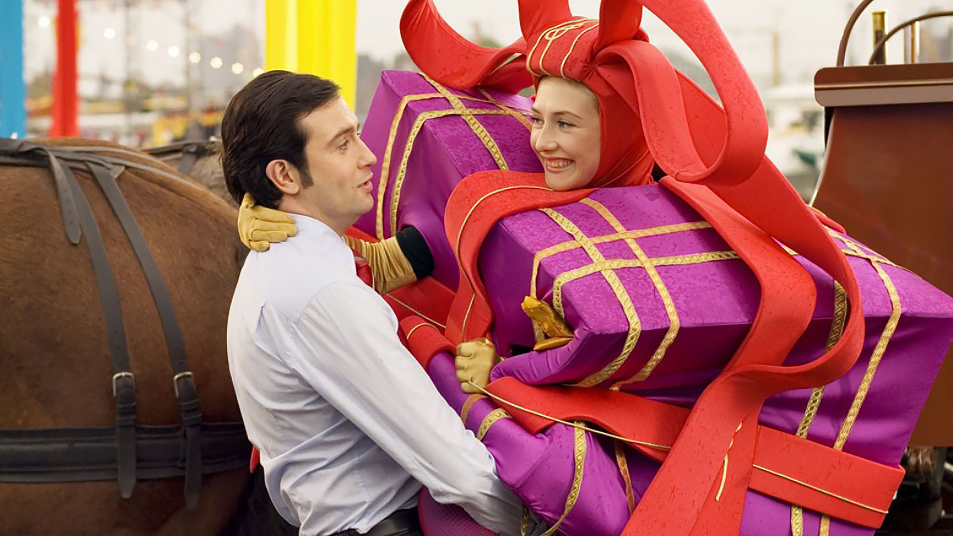 Een man in een wit overhemd houdt een vrouw vast die verkleed is als een groot, paars cadeau met rode strikken.