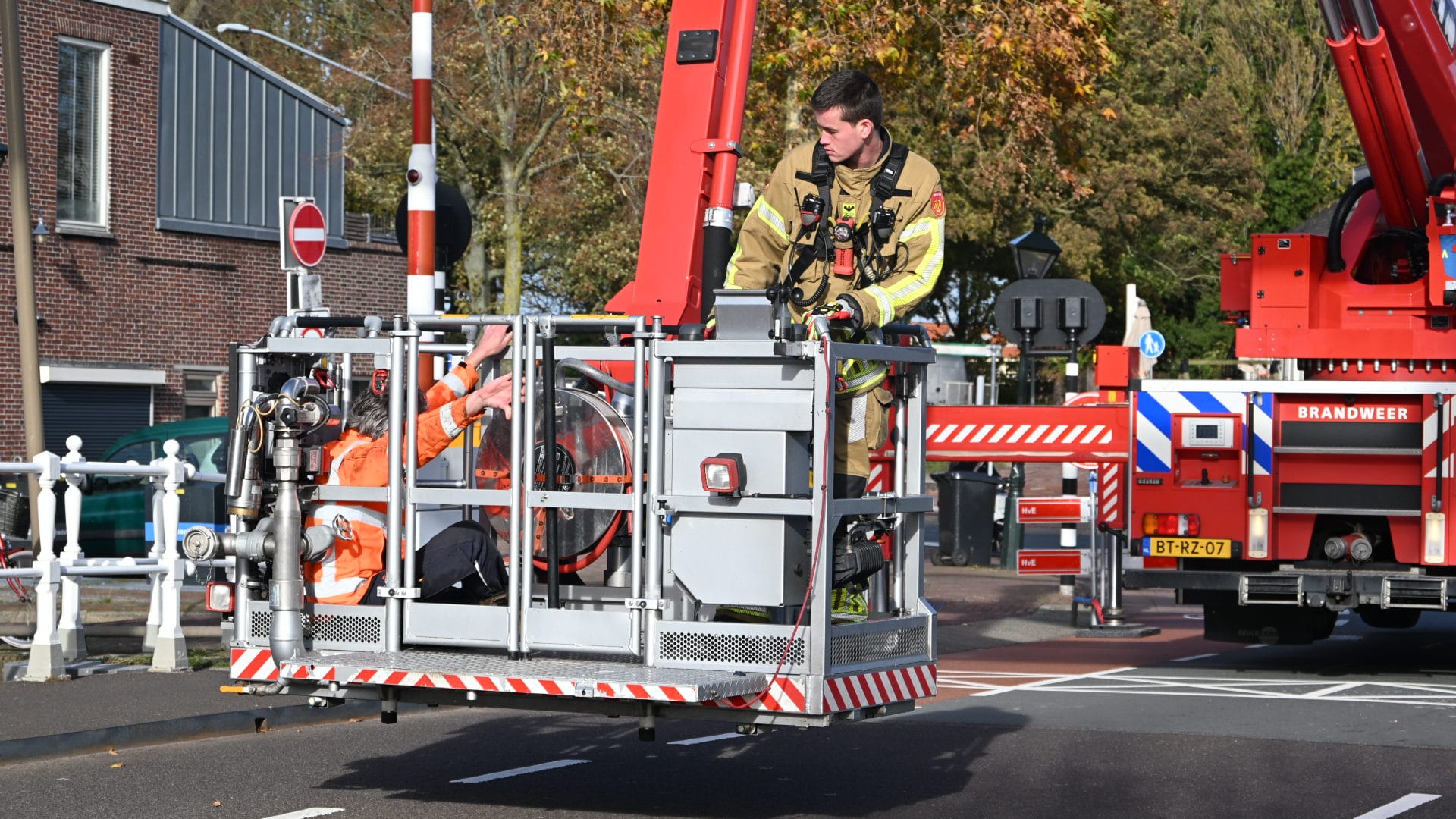 Een brandweerman in een kraanbak bestuurt een rode brandweerwagen op straat, met daarnaast een persoon in een oranje veiligheidsvest. De omgeving toont een stedelijke setting met bakstenen gebouwen en herfstbomen.