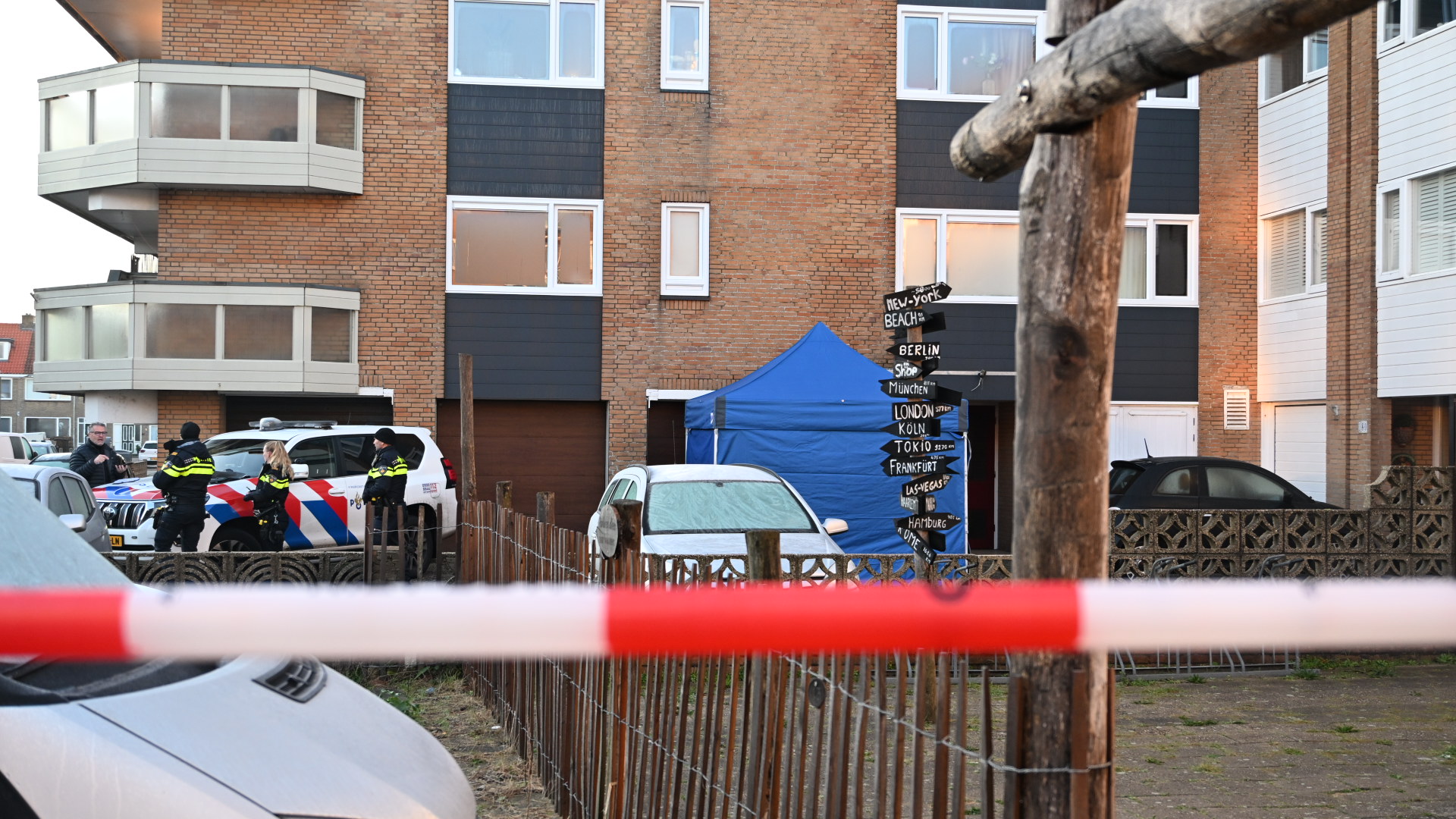 Politieagenten staan naast een politievoertuig voor een bakstenen gebouw met een blauwe tent. Een rood-wit afzetlint is op de voorgrond zichtbaar.