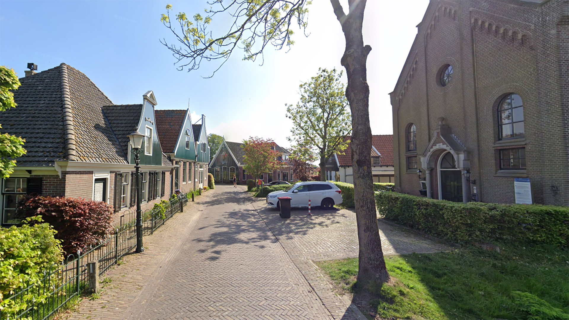Een straat in een Nederlands dorp met traditioneel vormgegeven huizen met puntgevels aan de linkerzijde en een groot bakstenen gebouw aan de rechterzijde. Er is een boom, een geparkeerde auto en een met struiken omheind gebied zichtbaar.