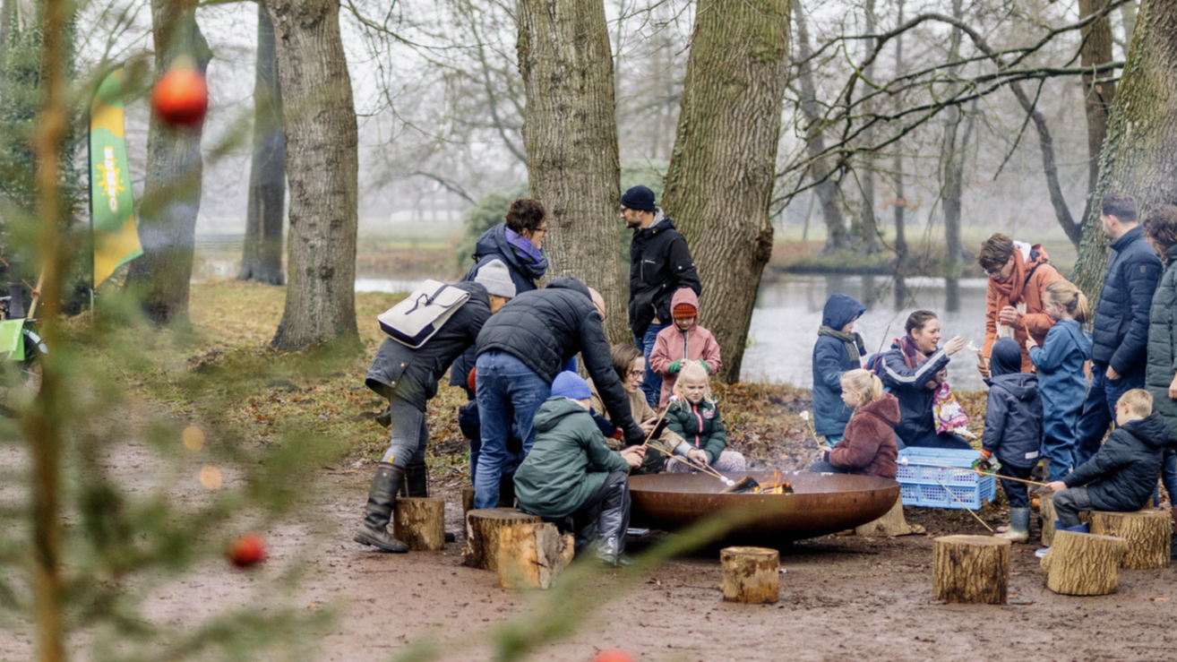 Een groep mensen, waaronder kinderen, zit rond een kampvuur in een bosrijke omgeving met bomen en een vijver op de achtergrond; op de voorgrond is een kerstboom met rode kerstballen zichtbaar.