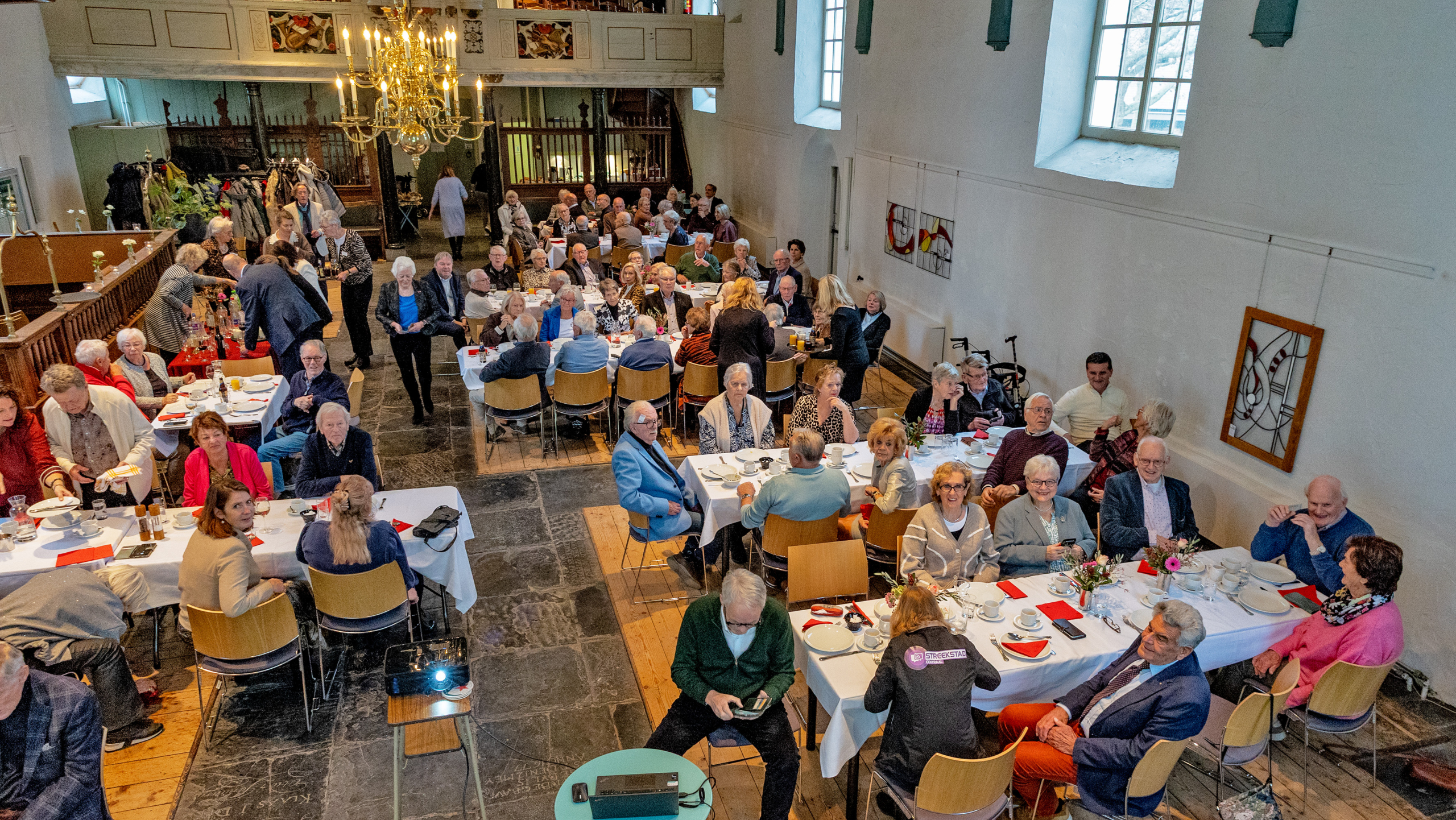 Groep mensen zit aan tafels in een kerkzaal tijdens een bijeenkomst, met witte tafelkleden en bloemen op de tafels en een kroonluchter bovenaan.