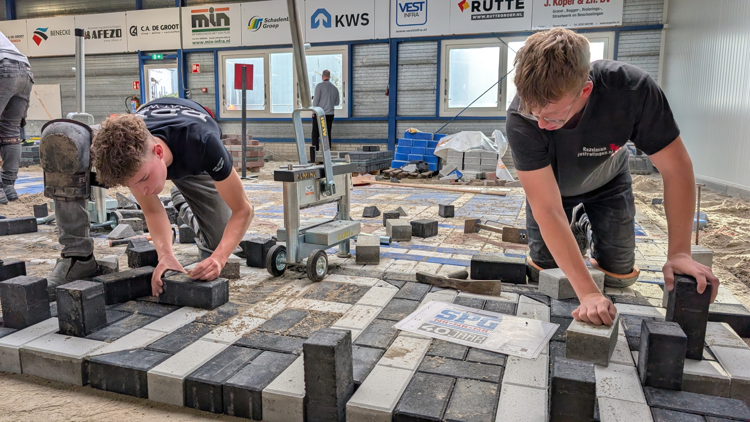 Twee jonge mannen met kniebeschermers leggen stenen in een patroon op de grond in een industriële ruimte, omringd door bouwmateriaal en reclameborden aan de muur.