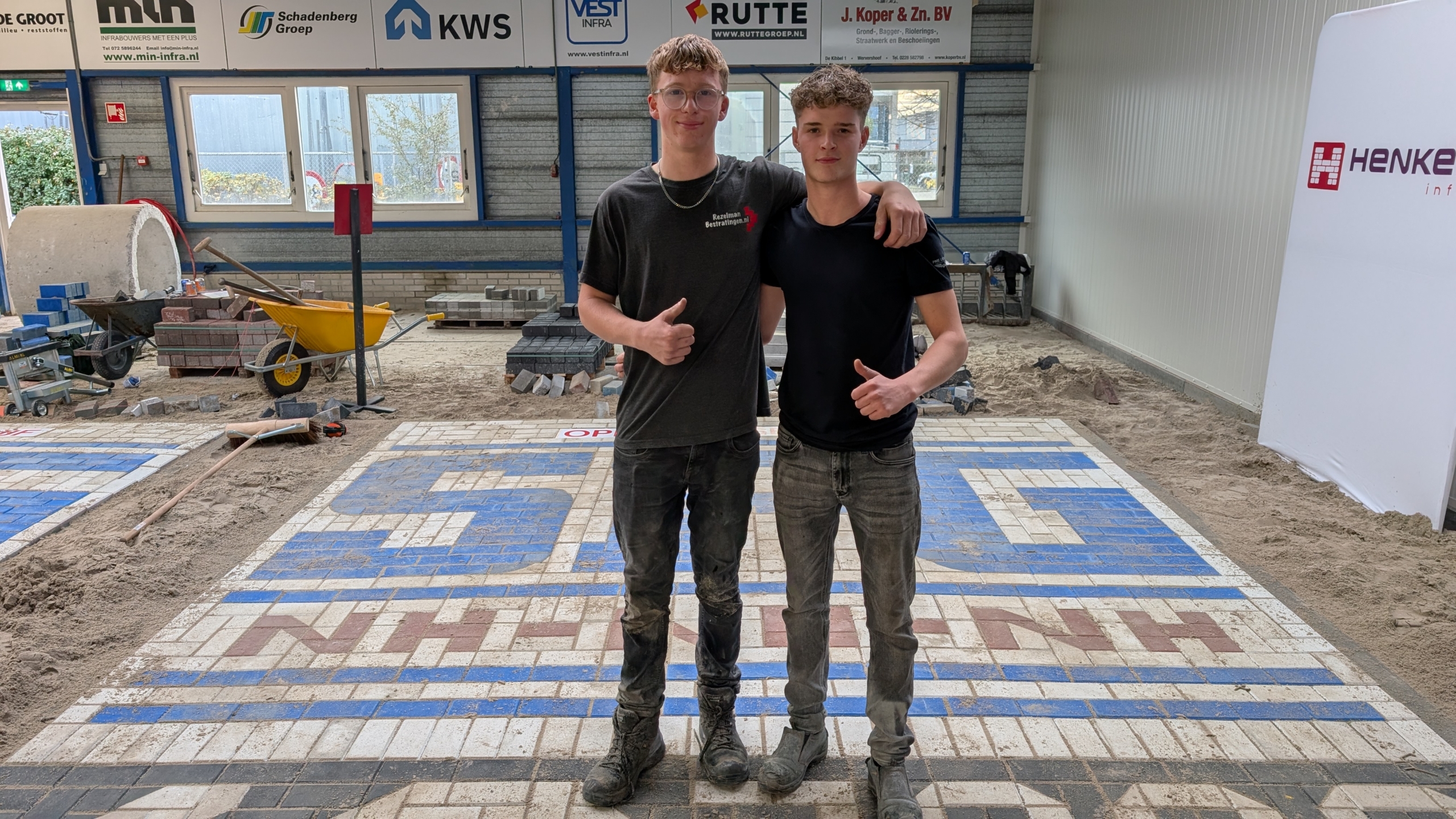 Twee jonge mannen staan zij aan zij met hun armen over elkaars schouders in een ruimte met bestrating in constructie. Ze geven allebei een duim omhoog en dragen werkkleding bedekt met stof. Op de achtergrond is bouwmateriaal zichtbaar, zoals een kruiwagen en stapels stenen, onder borden met bedrijfslogo's.