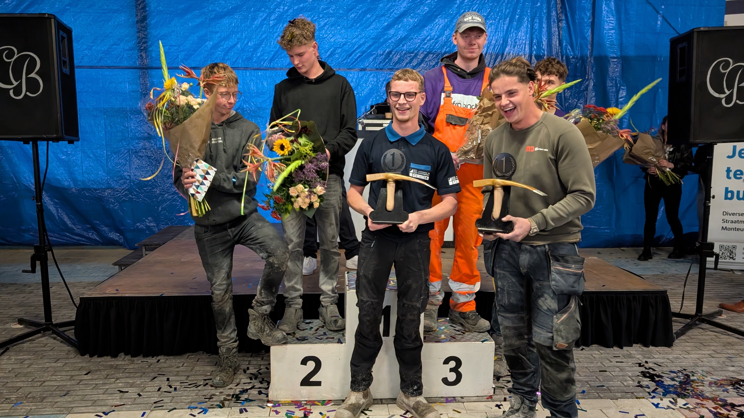 Een groep mannen poseert op een podium; twee van hen houden een prijs vast en anderen hebben bloemen in hun hand.