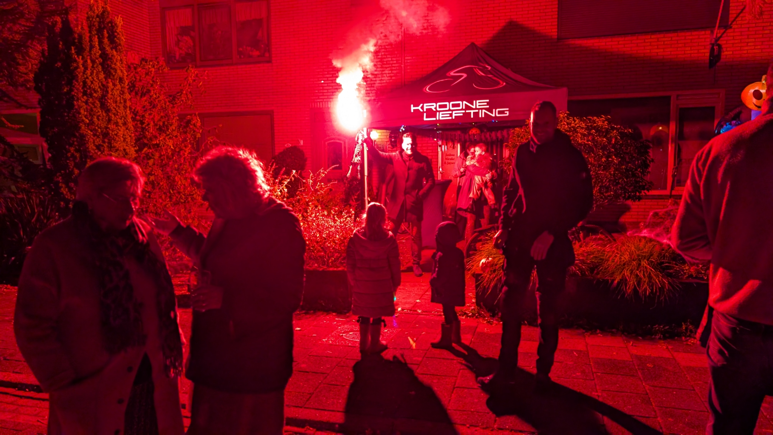 Nachtelijke scène met mensen voor een verlichte tent, waarbij een persoon een fakkel vasthoudt die een rood licht verspreidt.