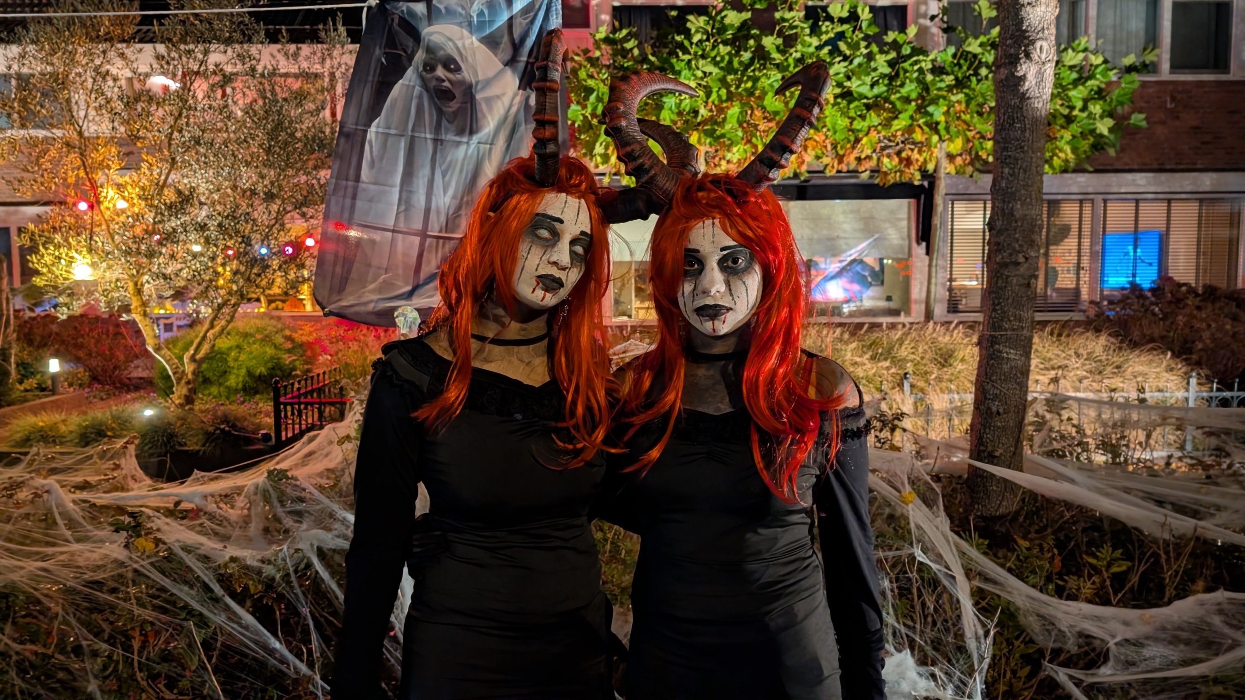 Twee mensen gekleed als demonische figuren met rode pruiken en grote hoorns dragen zwarte kleding; ze staan in een versierde tuin met spinnenwebben, met Halloween-verlichting en een spookachtige decoratie op de achtergrond.