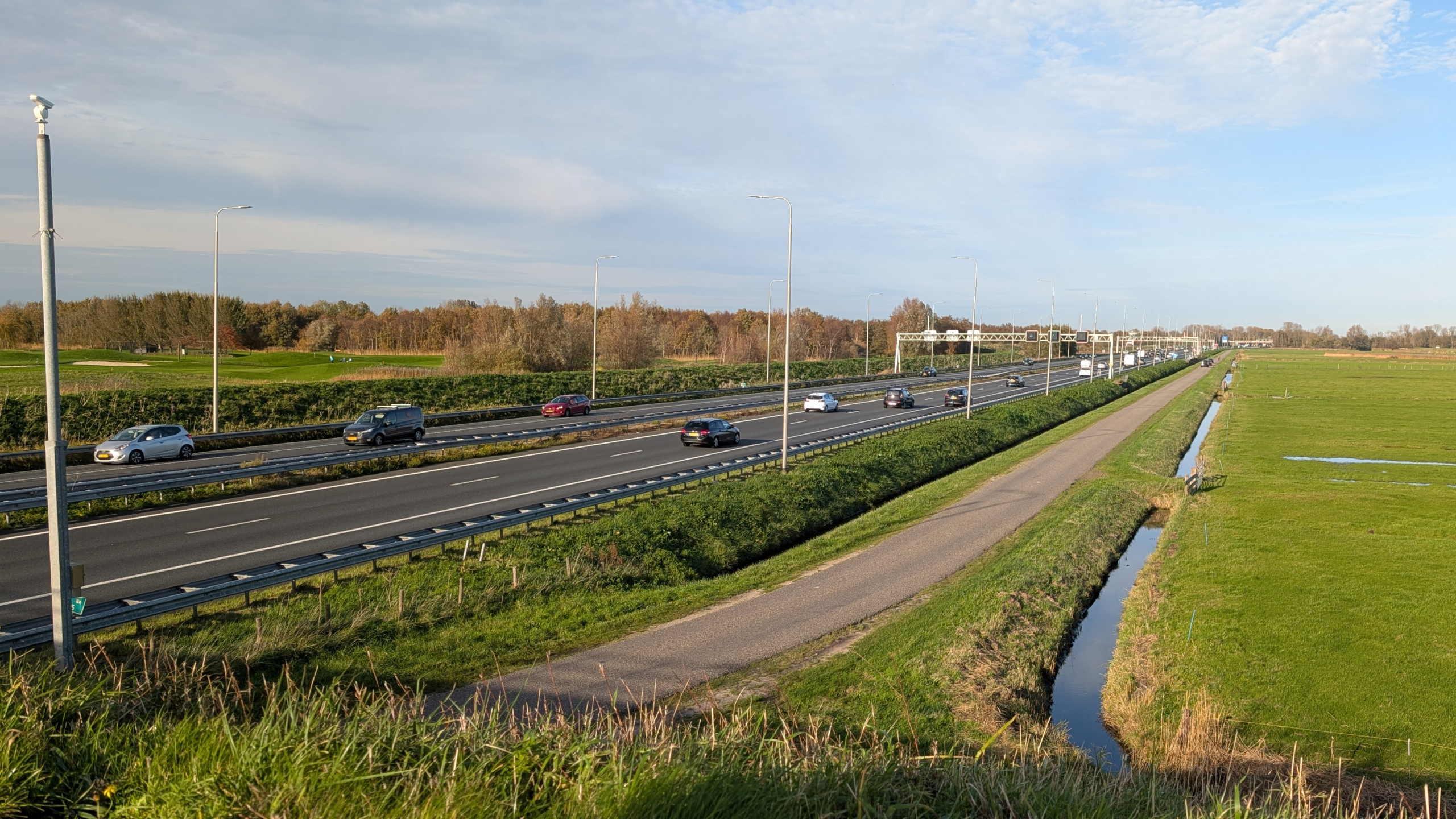 Drukke snelweg met auto's onder een hemel met lichte wolken, omgeven door groene velden en een smalle sloot; lantaarnpalen langs de weg.