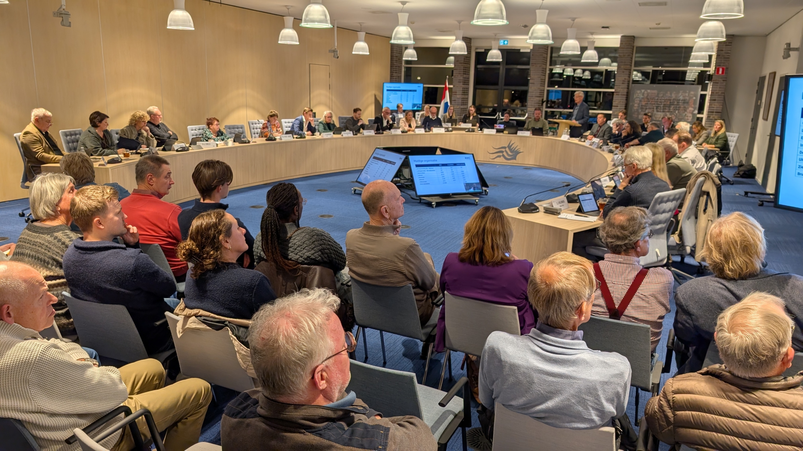 Een gemeenteraadsvergadering in een moderne zaal met deelnemers die rond een ovale tafel zitten, omringd door publiek dat op stoelen zit. Grootschermen tonen informatie, en de zaal heeft een professionele uitstraling met veel verlichting.