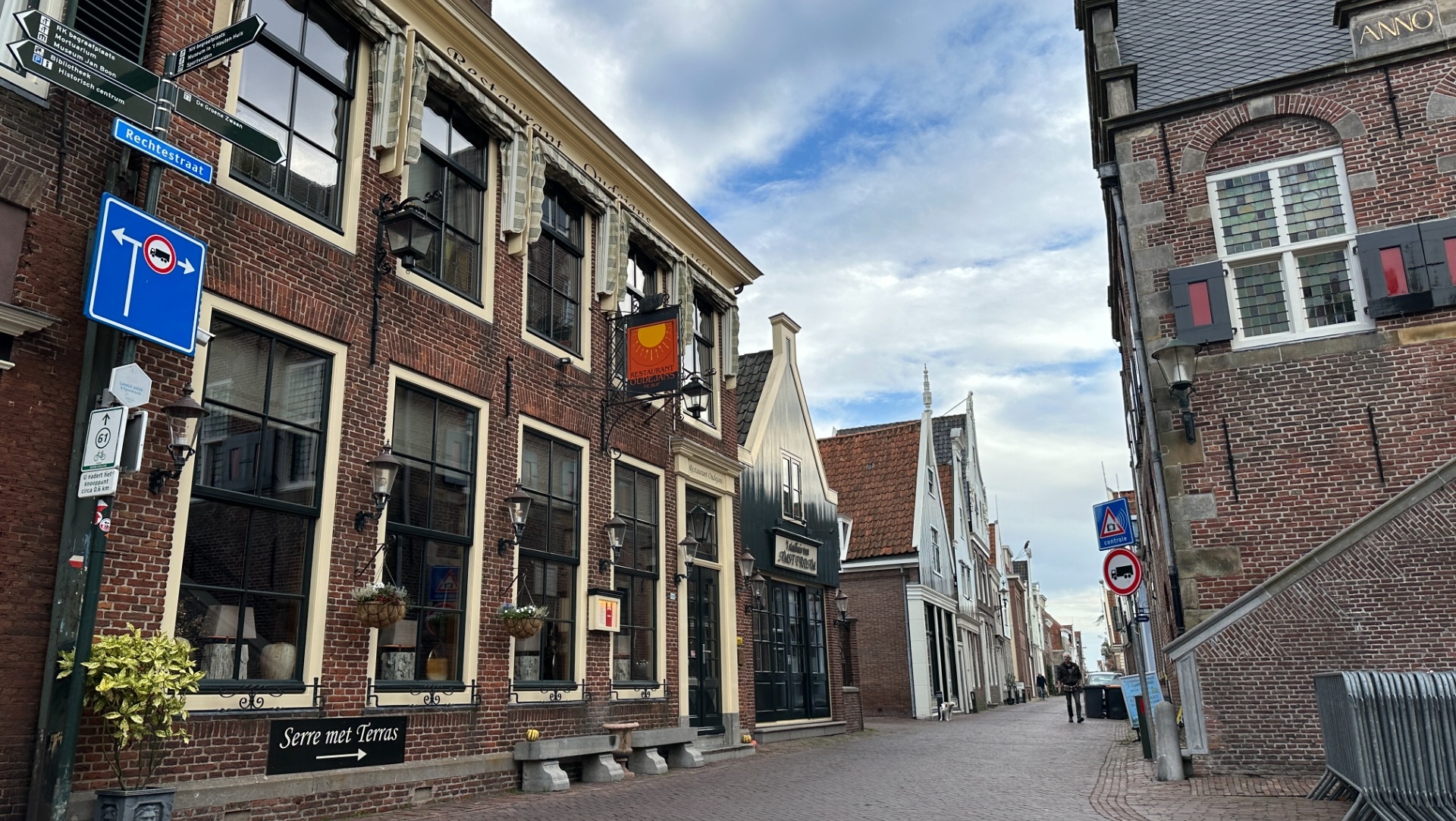Een rustige straat in een Nederlandse stad met historische bakstenen gebouwen aan weerszijden, verkeersborden op de voorgrond en een blauwe lucht met enkele wolken.