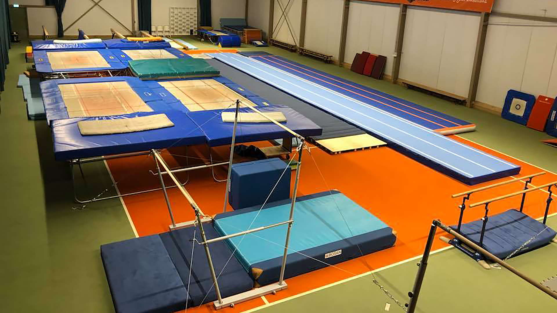 Een gymnastiekhal met diverse apparatuur, waaronder trampolines, een tumblingbaan en evenwichtsbalken, ligt op een oranje vloer met groene randen.