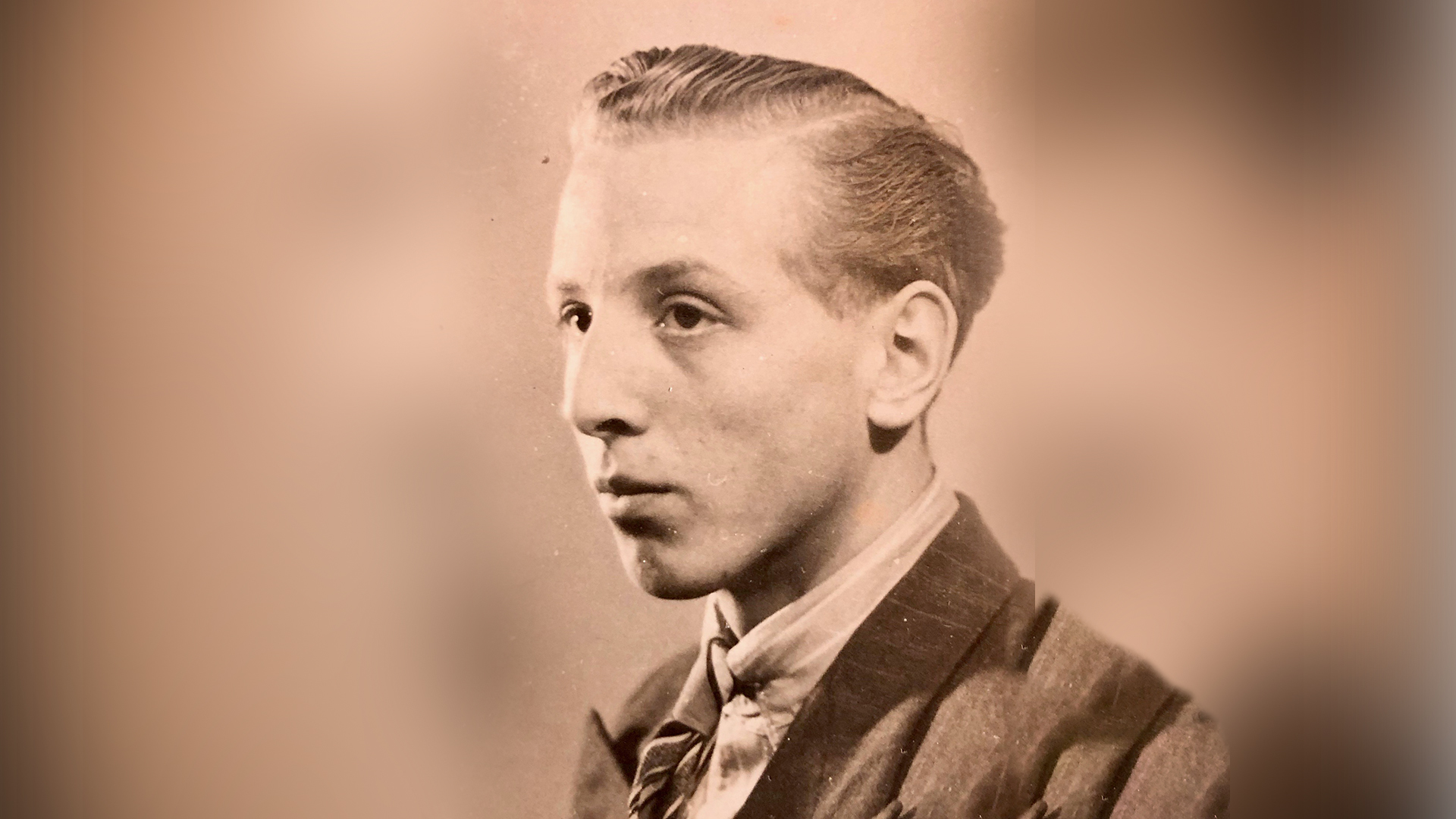 Zwart-wit portret van een jongeman in een pak met gestreepte das, met strak achterover gekamd haar, kijkend naar rechts.