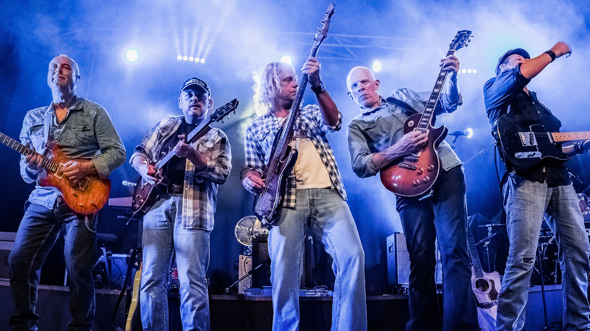 Een live-optreden van een band met vijf mannen die gitaren bespelen, in een sfeervolle belichting met rook op de achtergrond.