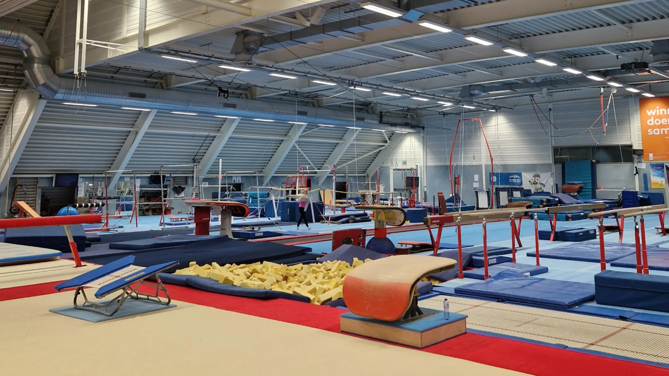 Binnenzicht van een gymnastiekhal met diverse oefeningenapparatuur zoals een springplank, een schuimrubberbak, ringen, balansbalken en matten.