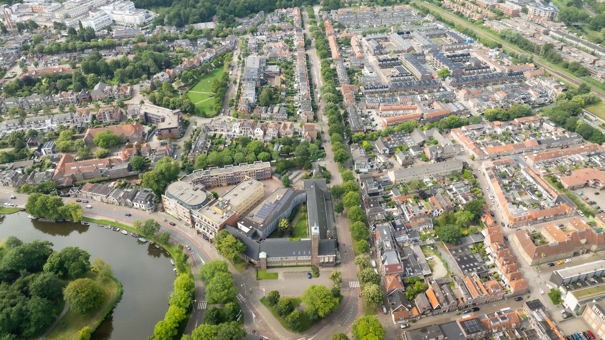 Luchtfoto van een stedelijk gebied met een mix van historische en moderne gebouwen, een kronkelende weg langs een waterlichaam, en groenrijke parken verspreid tussen de woonwijken.