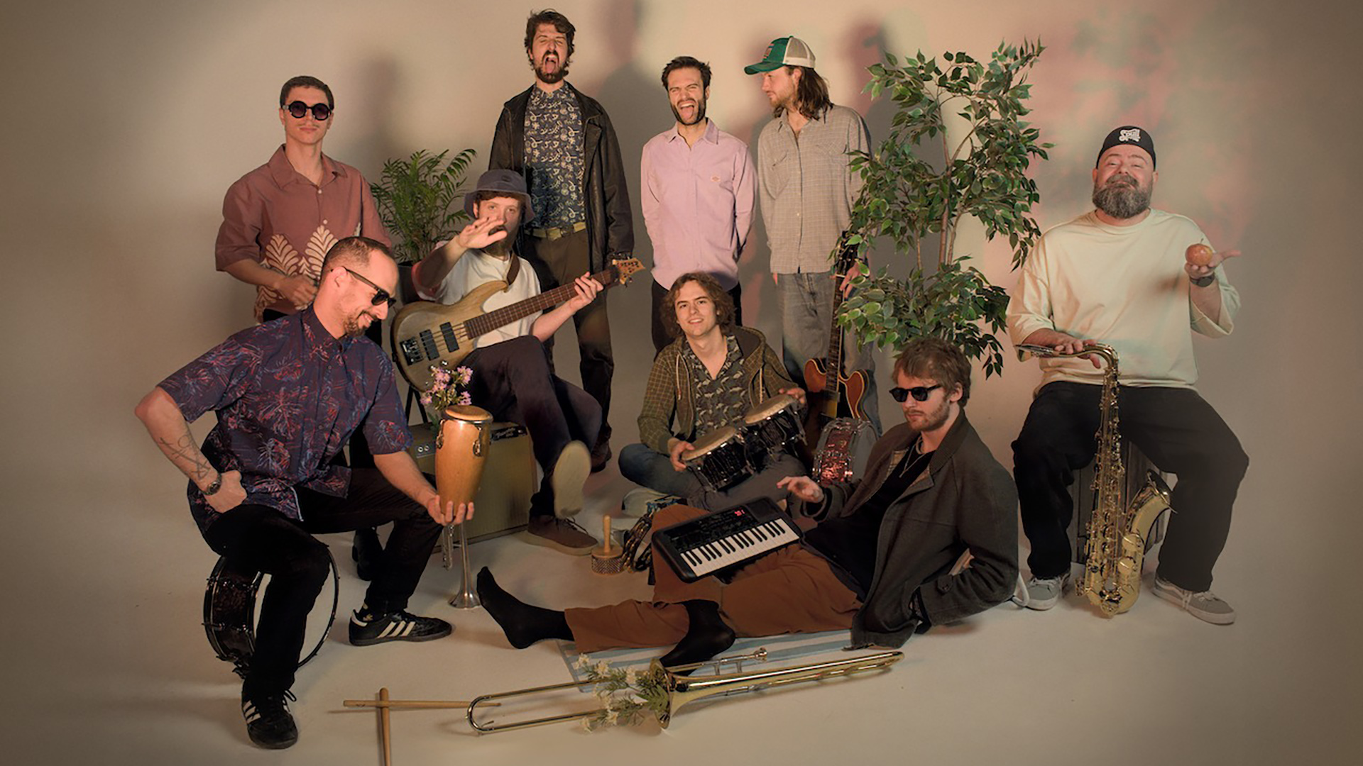 Een groep van negen muzikanten poseert met muziekinstrumenten en plantendecor op de achtergrond. Enkele muzikanten dragen zonnebrillen, terwijl een van hen een basgitaar vasthoudt en een ander een keyboard op schoot heeft. Er zijn ook percussie-instrumenten en een saxofoon zichtbaar.