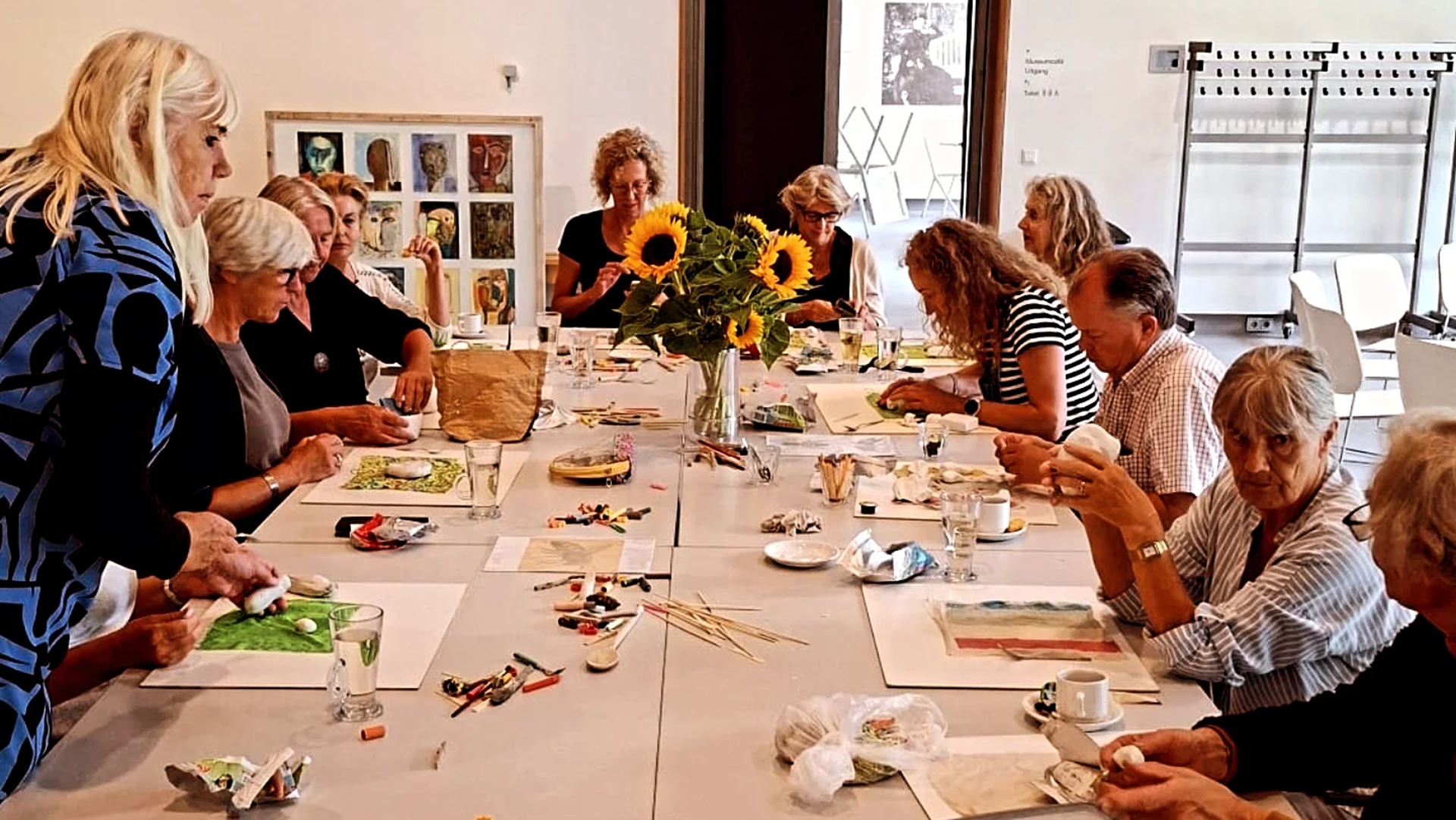 Een groep mensen zit rond een tafeltje met kunstbenodigdheden en bloemen in het midden, bezig met een kunstproject of workshop in een lichte ruimte.