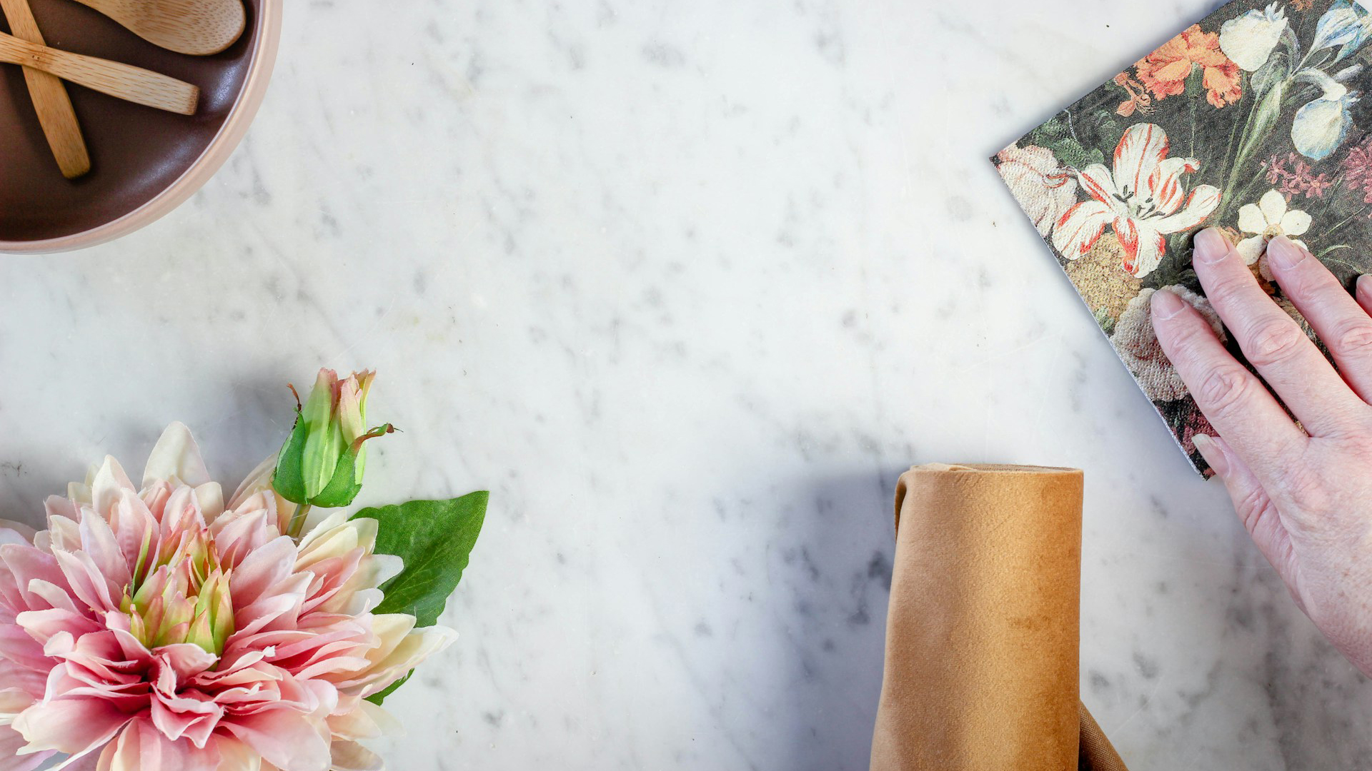 Bovenaanzicht van een marmeren tafel met een hand die een boek met bloemenachtergrond vasthoudt, een roze bloem met groene bladeren links, een bruine stoffen servet rechtsonder en een bruine kom met houten lepels linksboven.