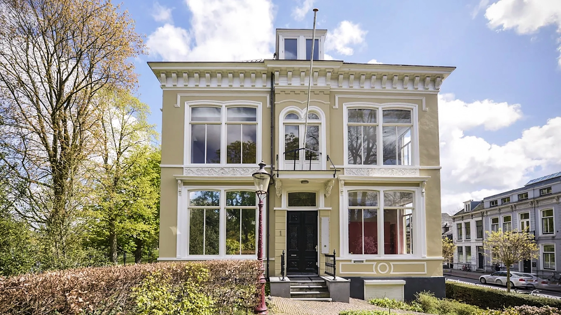 Groot, klassiek pand met twee verdiepingen en een symmetrische voorgevel, gelegen aan een laan met bomen, onder een heldere hemel met wolken.