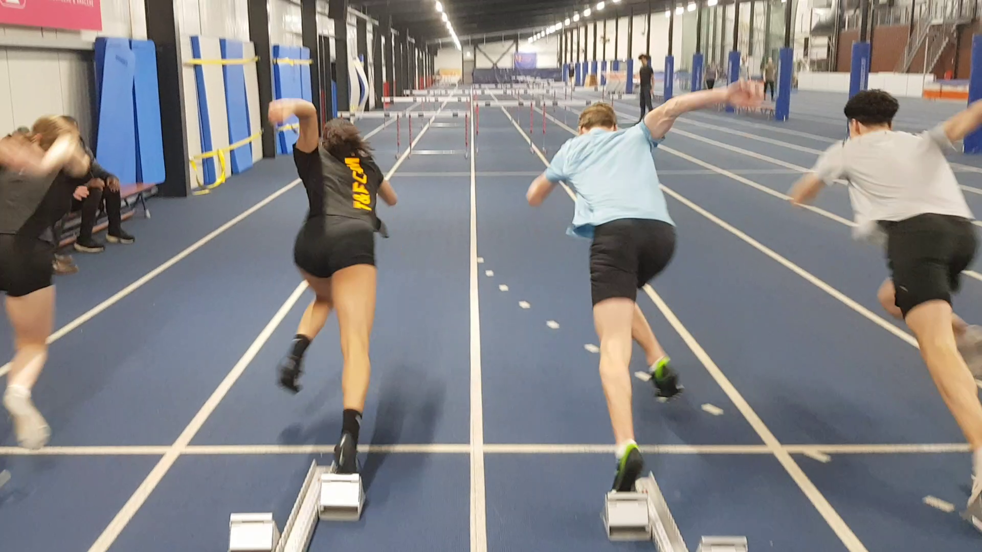 Vier atleten starten met rennen op een indoor atletiekbaan, met hordes op de baan zichtbaar verderop.