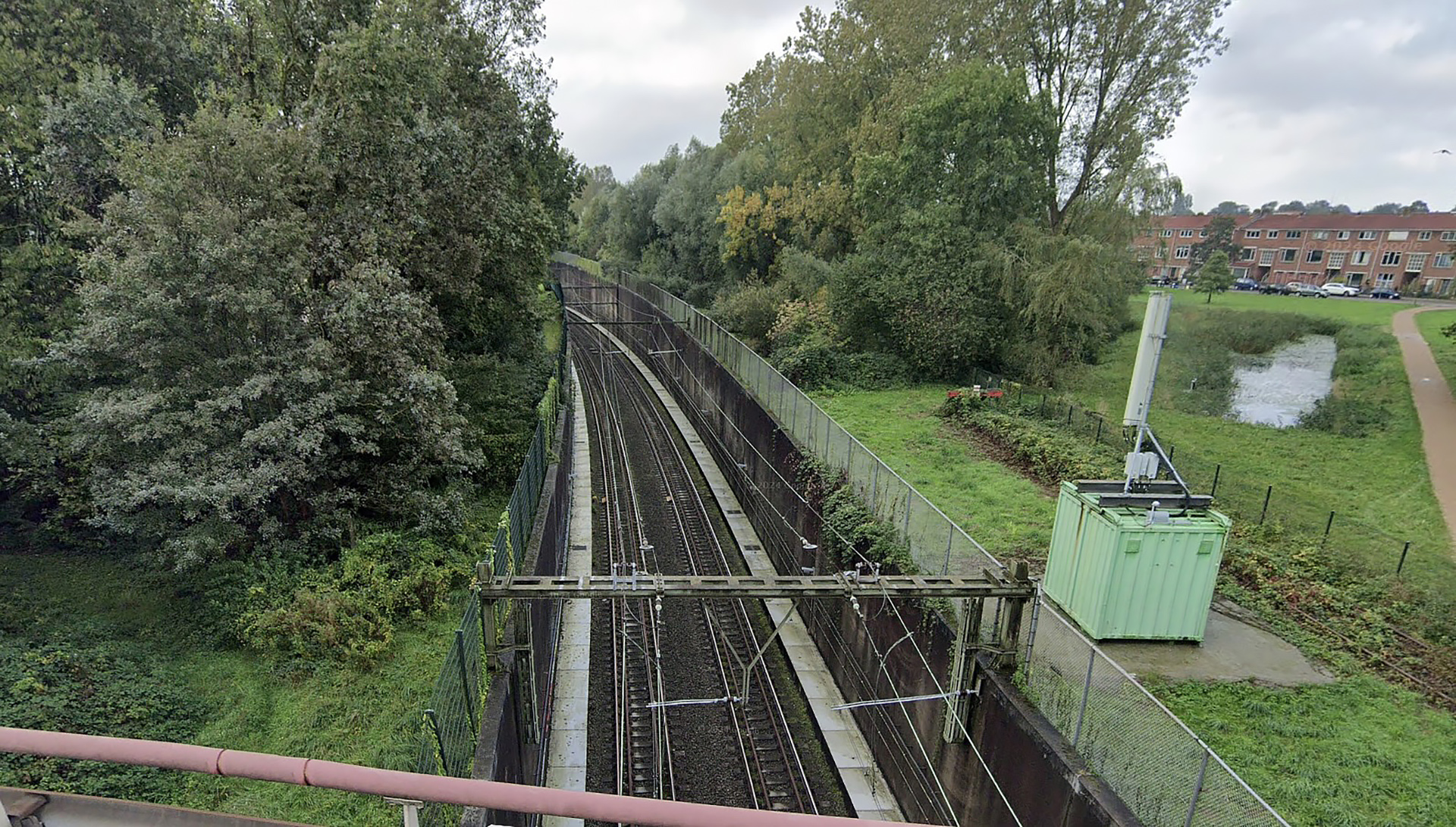 Treinsporen omgeven door groen, gezien vanaf een viaduct met links bomen en rechts een grasveld met een groene container.