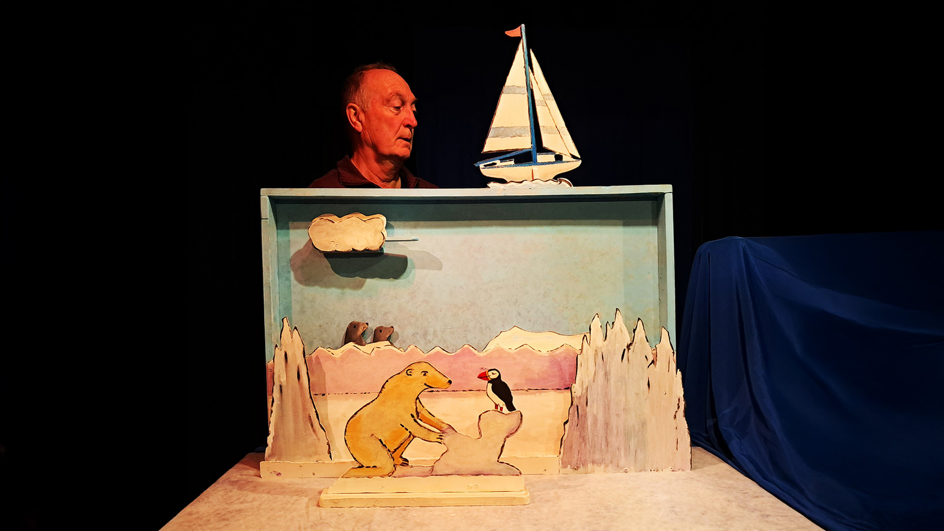 Man operates a small puppet theater depicting a polar bear and a puffin with a painted backdrop of icebergs and a sailboat.