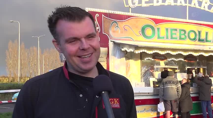 Man bij oliebollenkraam met een microfoon, kleurrijke kraam op de achtergrond met mensen die in de rij staan.