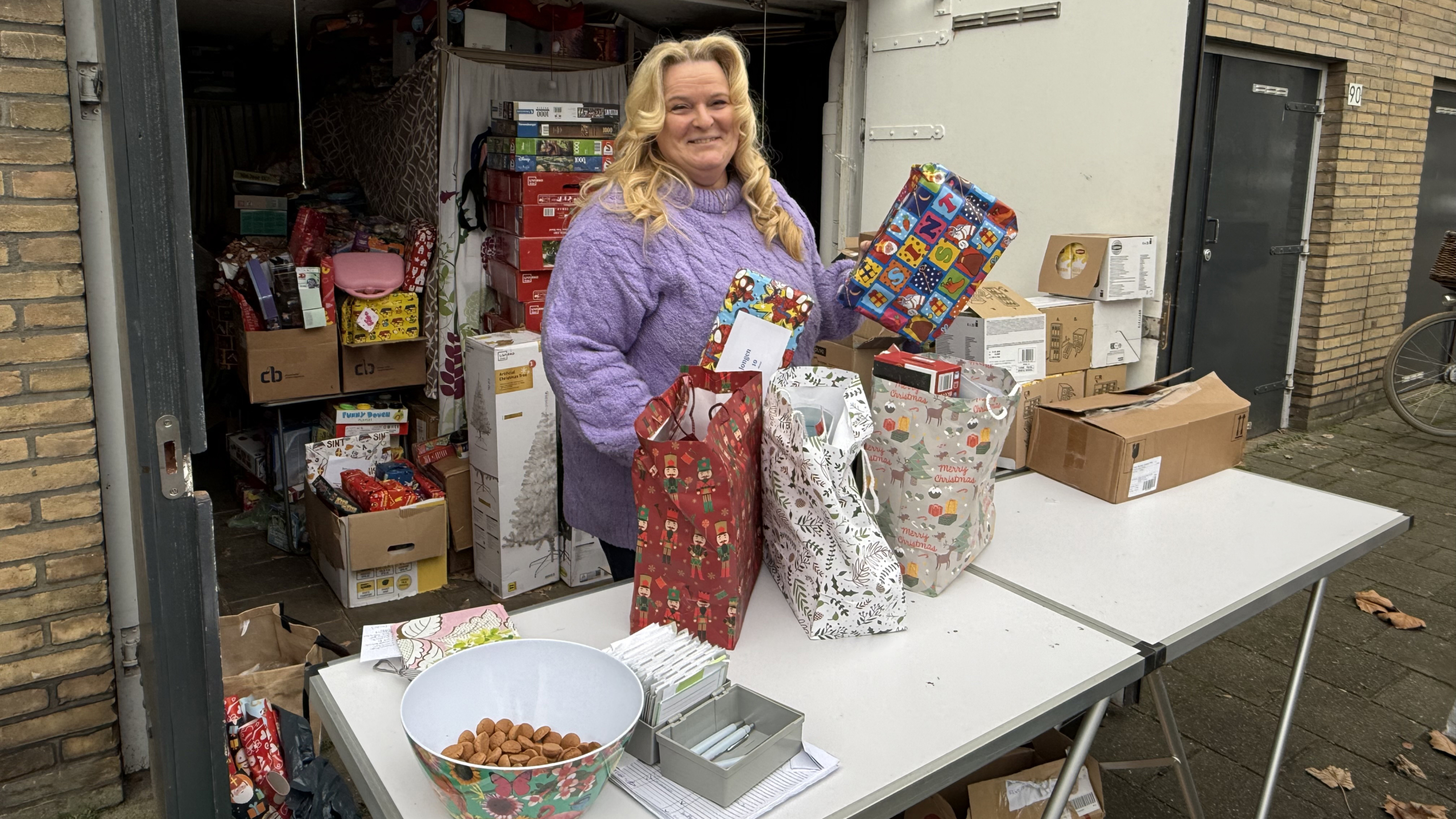 Vrouw in paarse trui toont ingepakt cadeau voor opslagruimte met cadeaus; tafel met noten en cadeautassen.