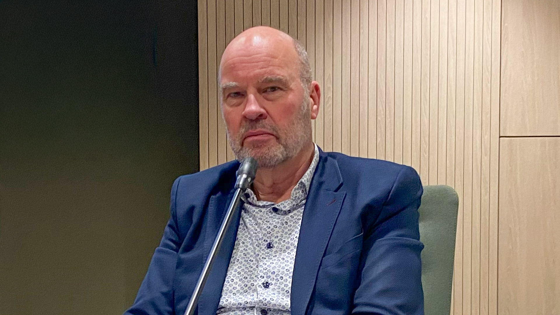 Een kalende man met een grijze baard, gekleed in een donkerblauw colbert en een overhemd met een patroon, zit voor een microfoon tegen een achtergrond van houten panelen.