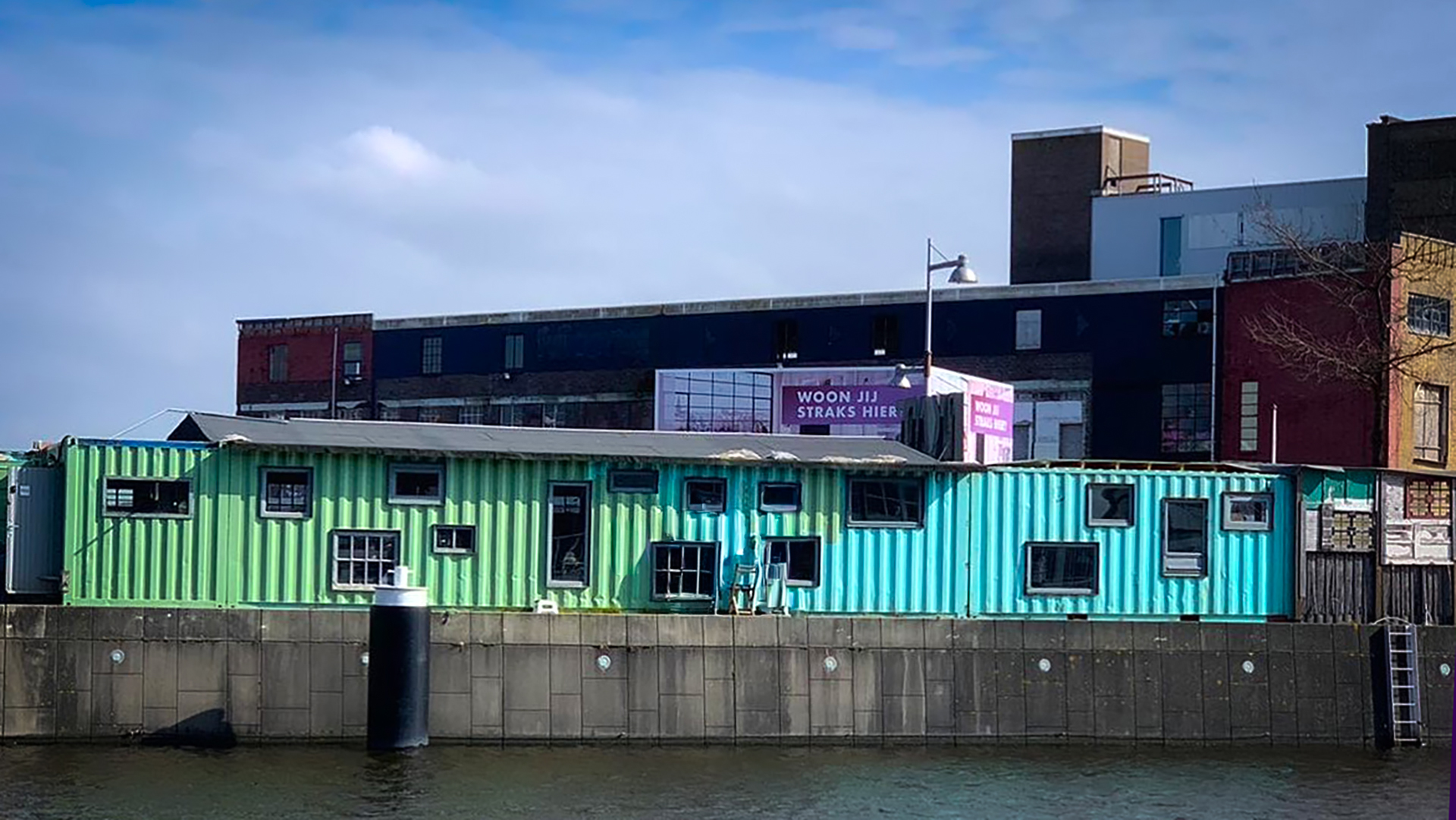 Een kleurrijk huisje gemaakt van zeecontainers aan het water, met daarachter een bakstenen gebouw en een bord met de tekst 'Woon jij straks hier'.