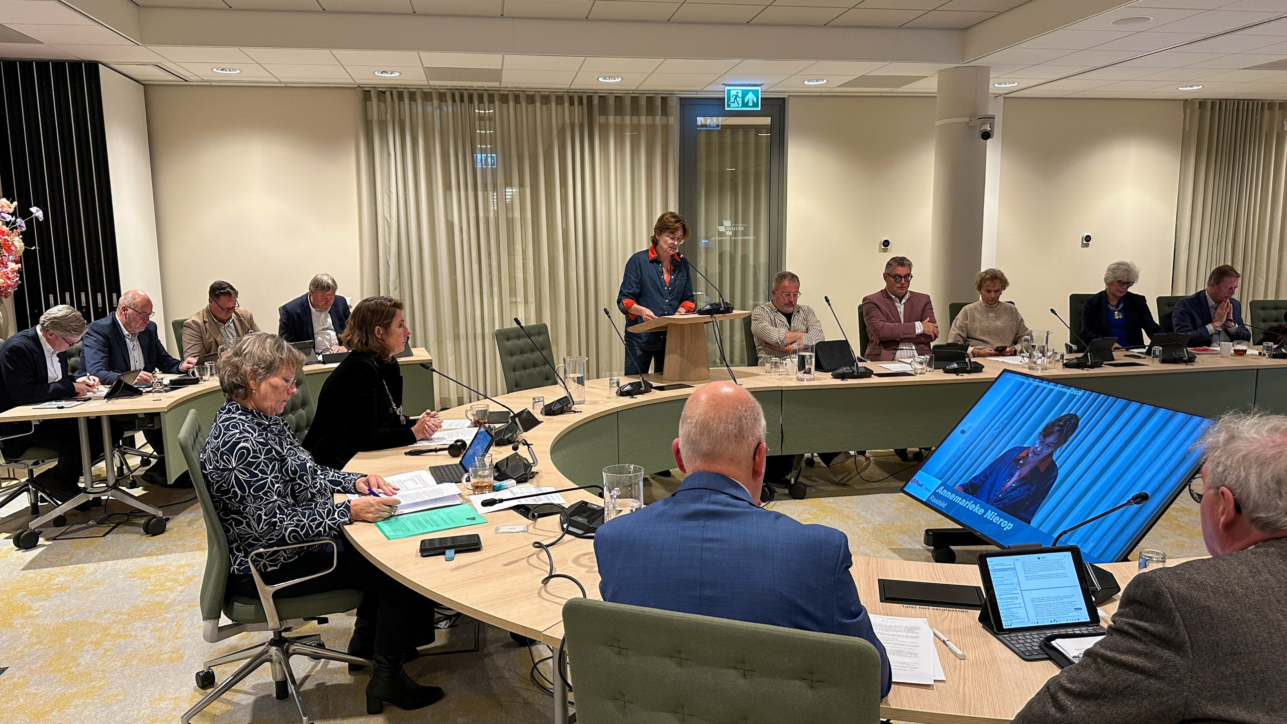 Een vergadering in een conferentiezaal met mensen die rond een ovale tafel zitten. Een vrouw staat achter een spreekgestoelte en spreekt in een microfoon, terwijl andere deelnemers aantekeningen maken of naar een scherm kijken.