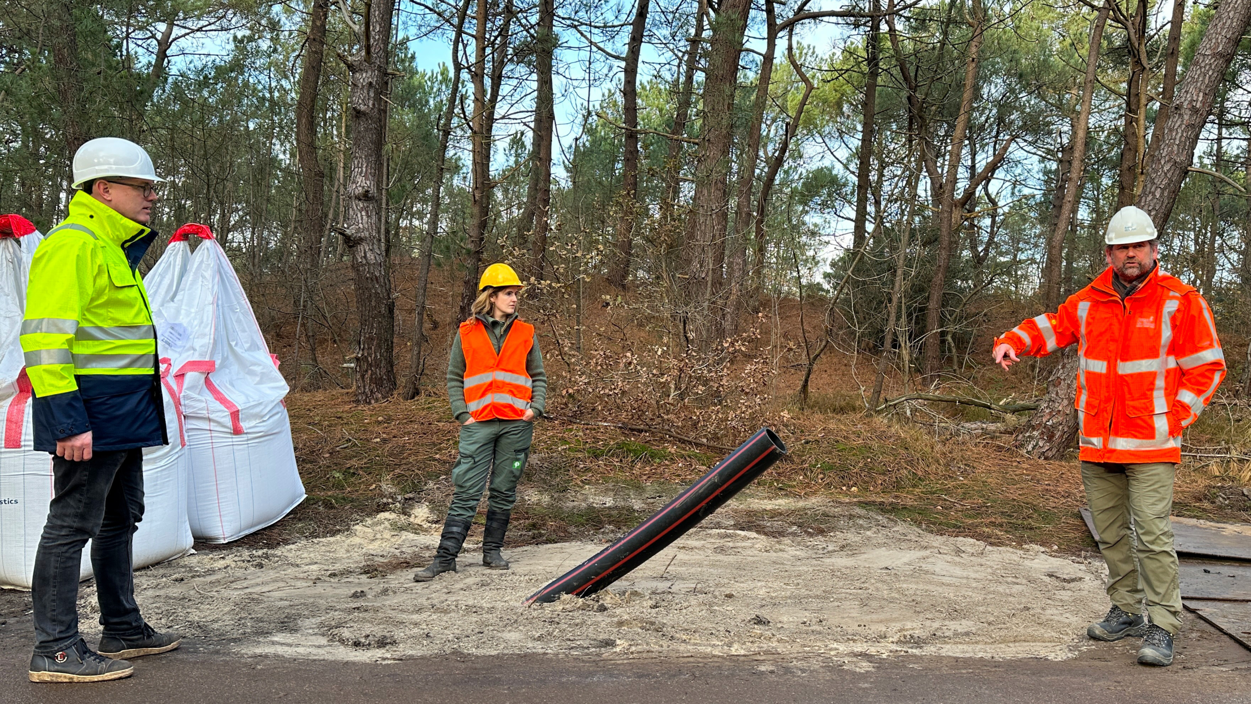 Drie mensen in veiligheidskleding en helmen staan bij een bouwplaats in een bosrijke omgeving, met een schuinstaande buis in de grond.