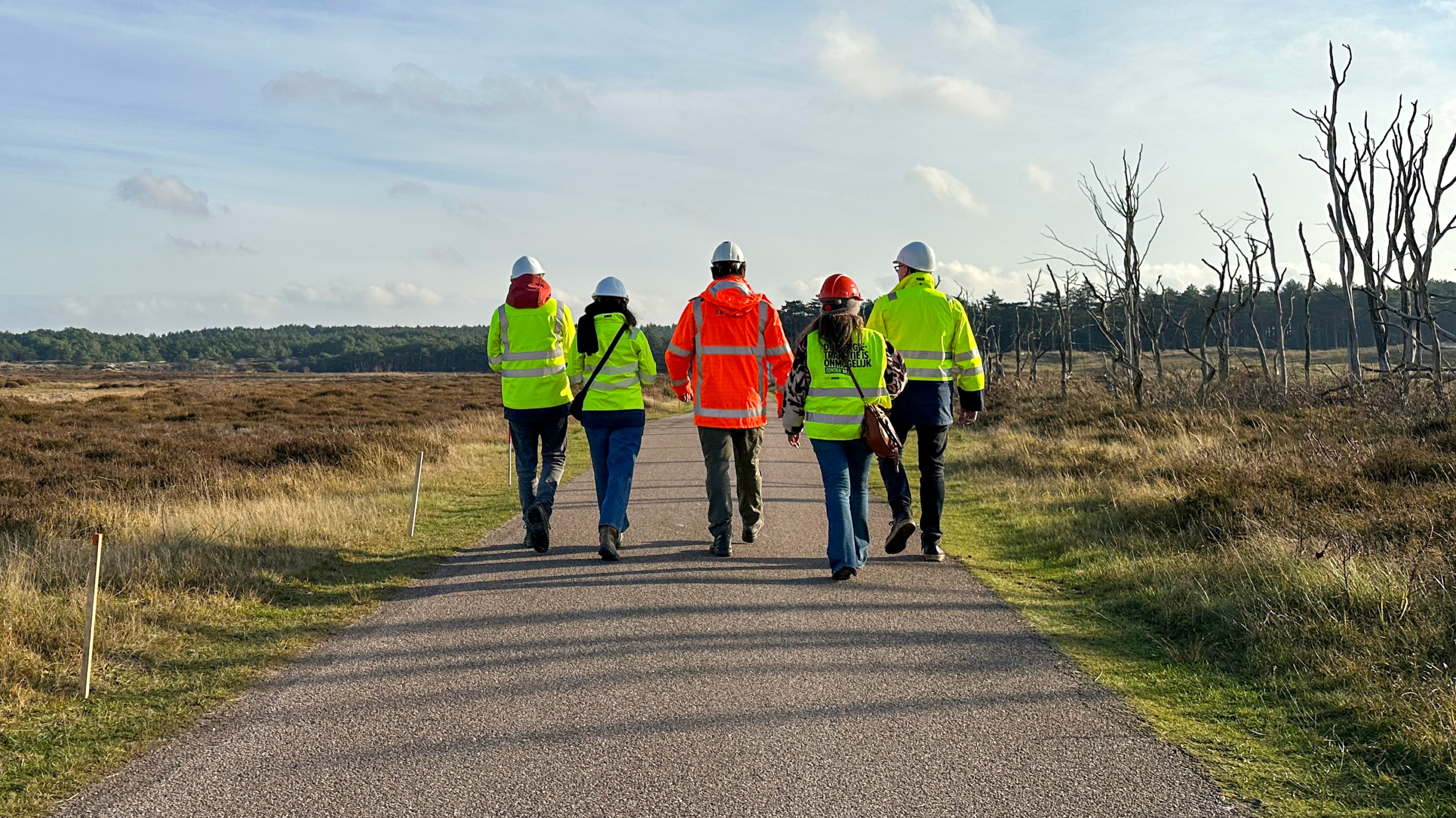 Vijf mensen in veiligheidshesjes en helmen lopen over een landelijk pad omringd door gras en bomen.