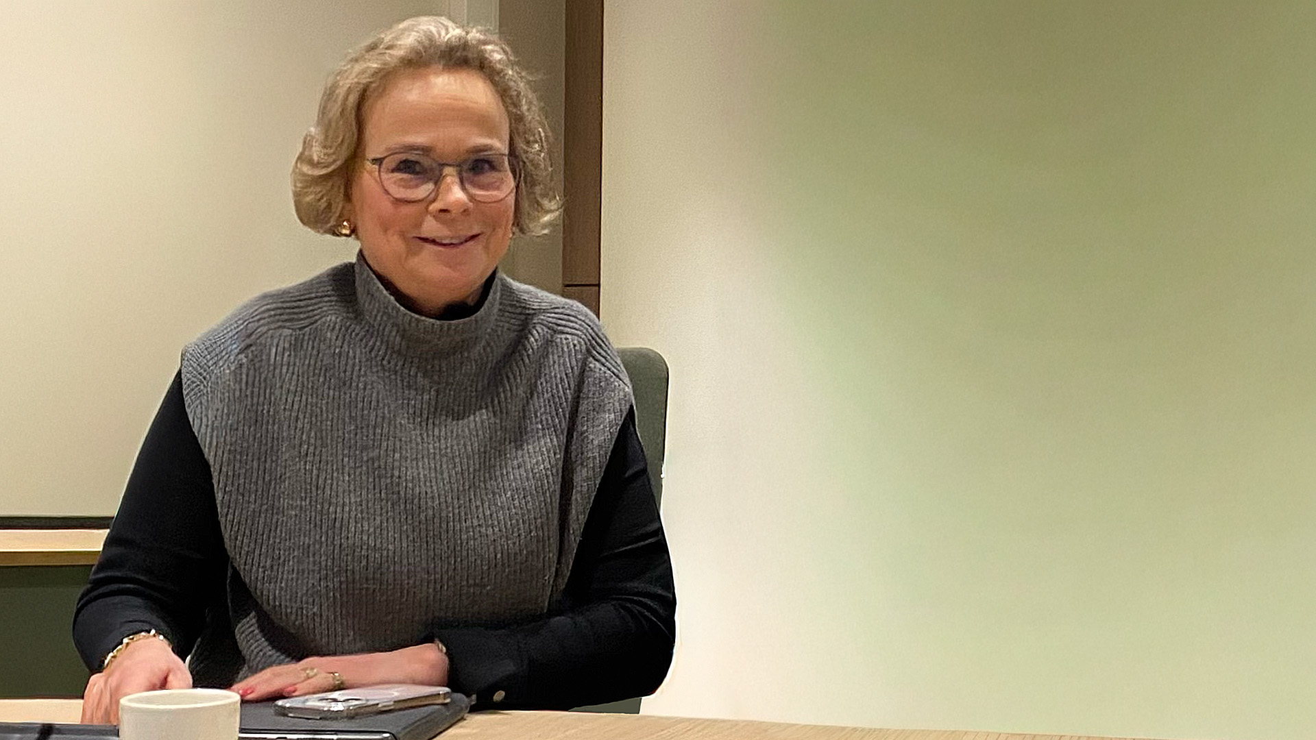 Vrouw met bril en grijs vest zit aan een tafel in een kantooromgeving.