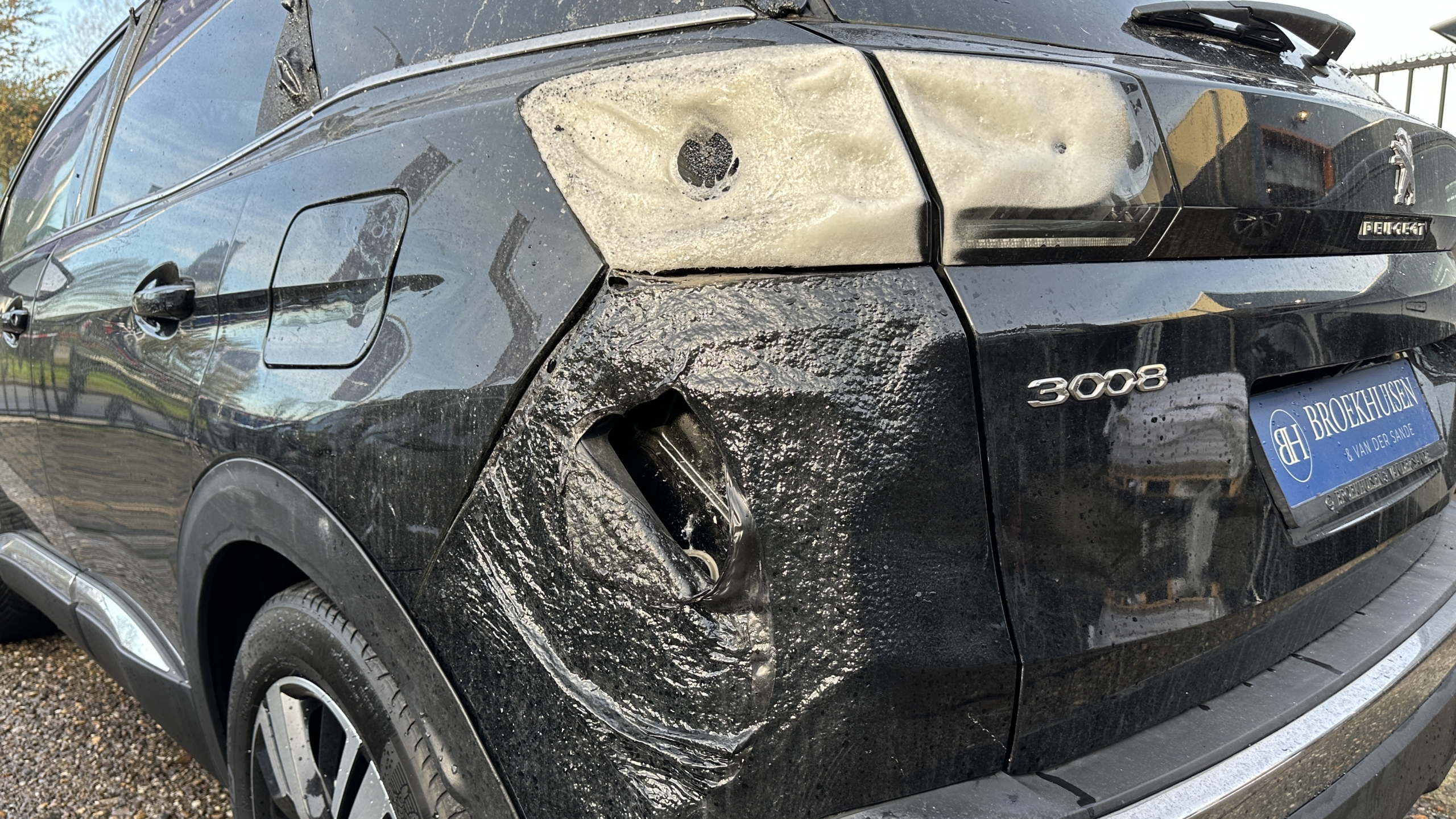 Een zwarte Peugeot 3008 met zichtbare brandschade aan de achterkant en zijkant, geparkeerd op een grindoppervlak voor een hek.