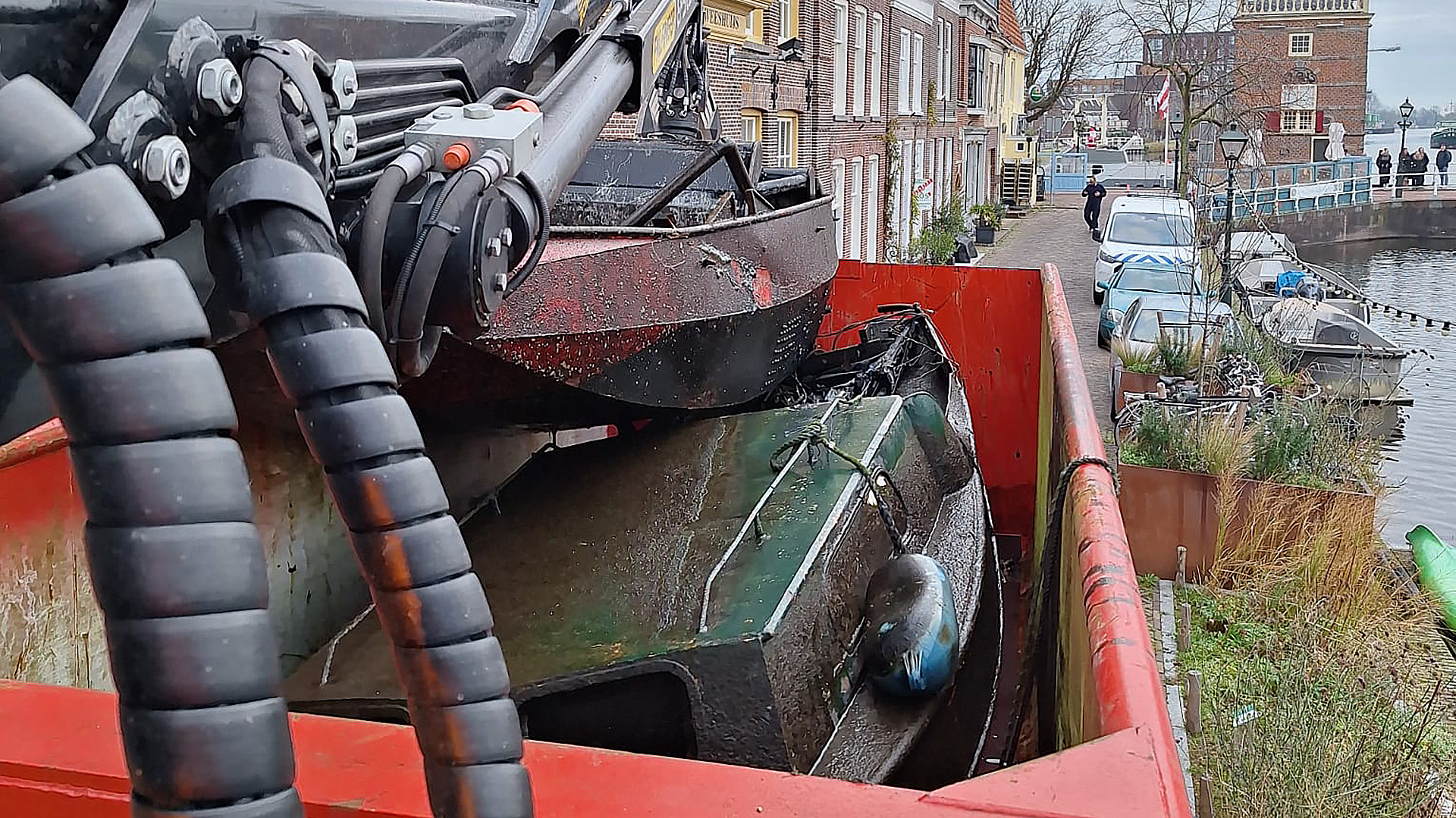 Een kraan tilt een klein, vuil schip uit het water en plaatst het in een rode bak in een smalle straat naast een kanaal met geparkeerde auto's en boten, omringd door bakstenen gebouwen.