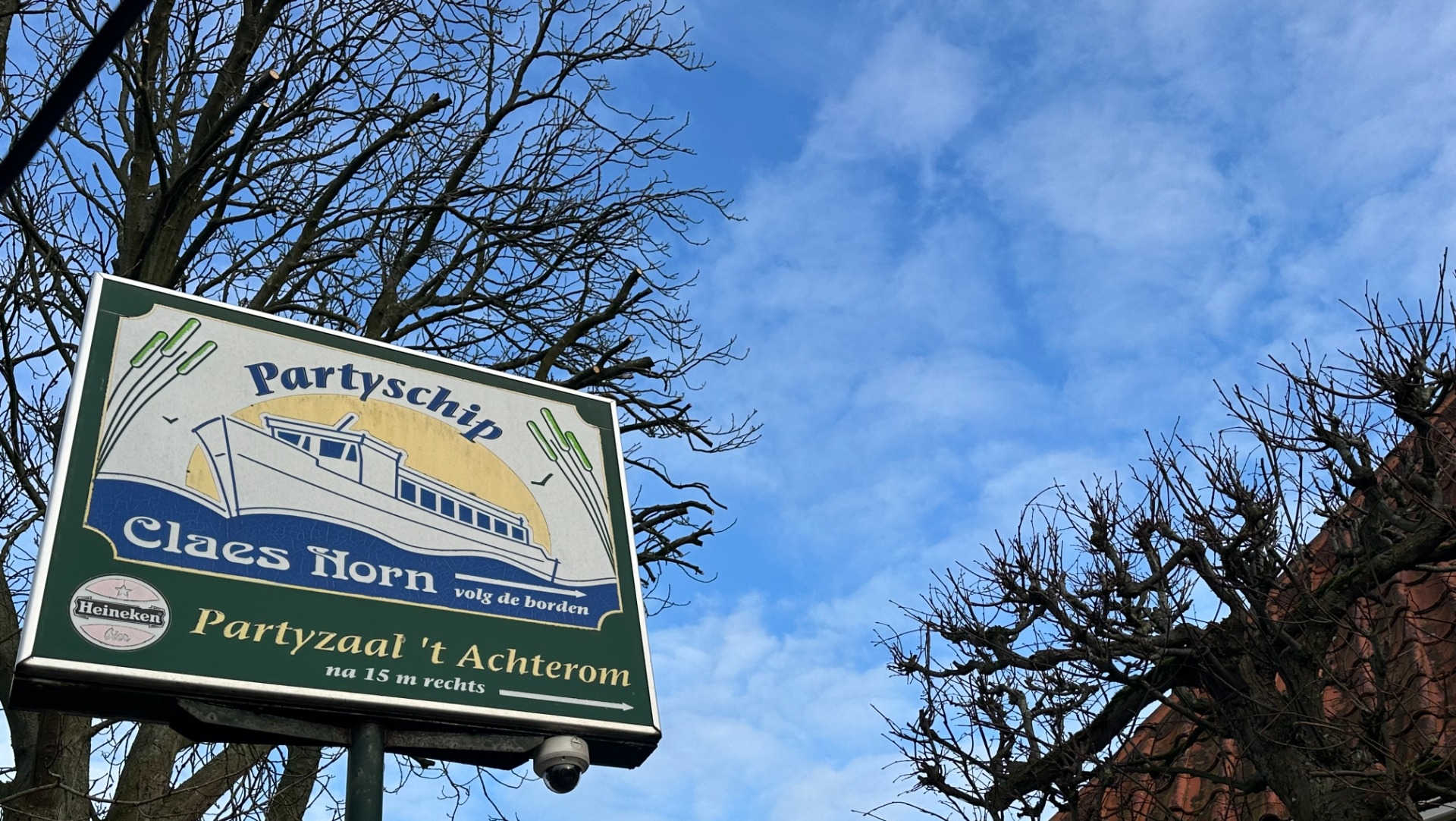 Bord met de tekst "Partyschip Claes Horn" op een paal, met een tekening van een boot erop, tegen een achtergrond van een boom en een blauwe lucht.