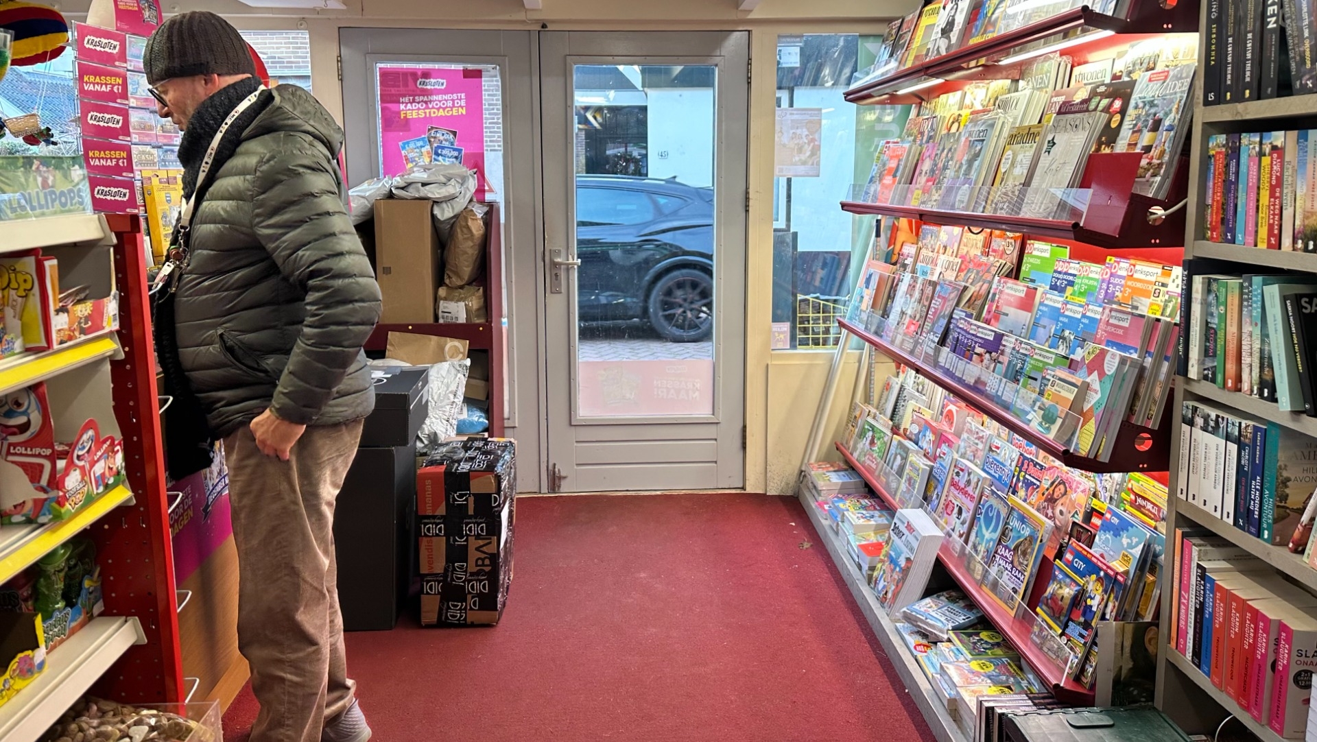 Een man in een warme jas en muts staat in een kleine winkel met tijdschriften en boeken aan de rechterkant en snoepgoed aan de linkerkant.