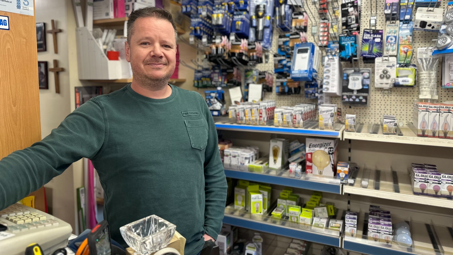 Man in een groene trui staat in een winkel met een assortiment aan huishoudelijke producten op de achtergrond.