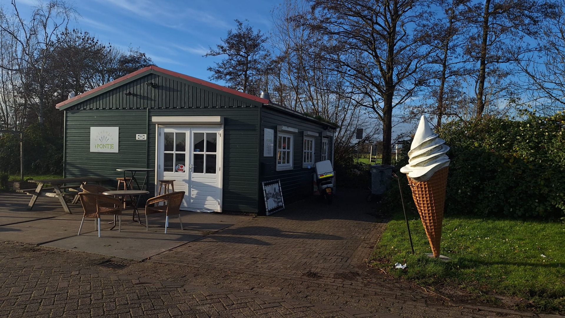 Een groene houten kiosk met een rode rand, omringd door bomen en struiken, met zitgelegenheid buiten. Links staat een groot ijsjesbeeld op het gras.