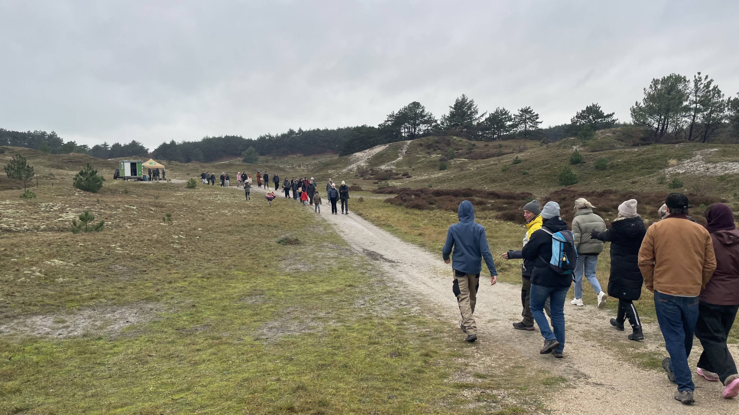 Groep mensen die over een wandelpad door een groen landschap met heuvels en bomen loopt, onder een bewolkte hemel.
