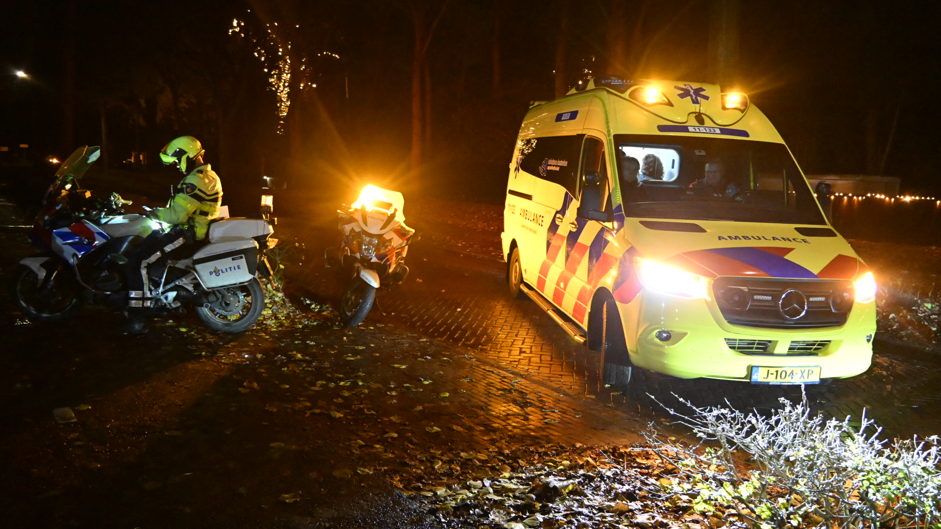 Een politieagent op een motor en een ambulance staan geparkeerd op een weg bedekt met herfstbladeren. Beide voertuigen hebben hun lichten aan in het donker.