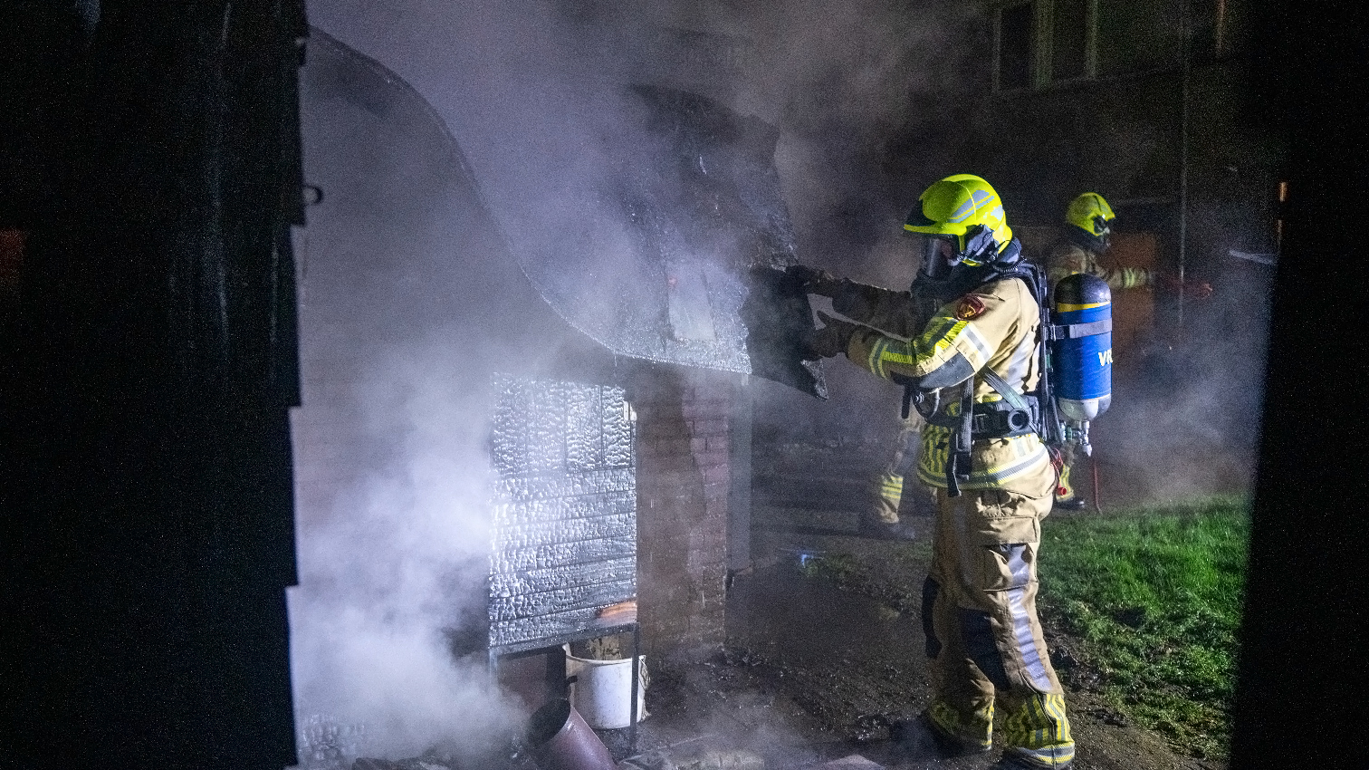 Brandweermannen in beschermende uitrusting en helmen blussen een brand buiten in de nacht met rook om hen heen.